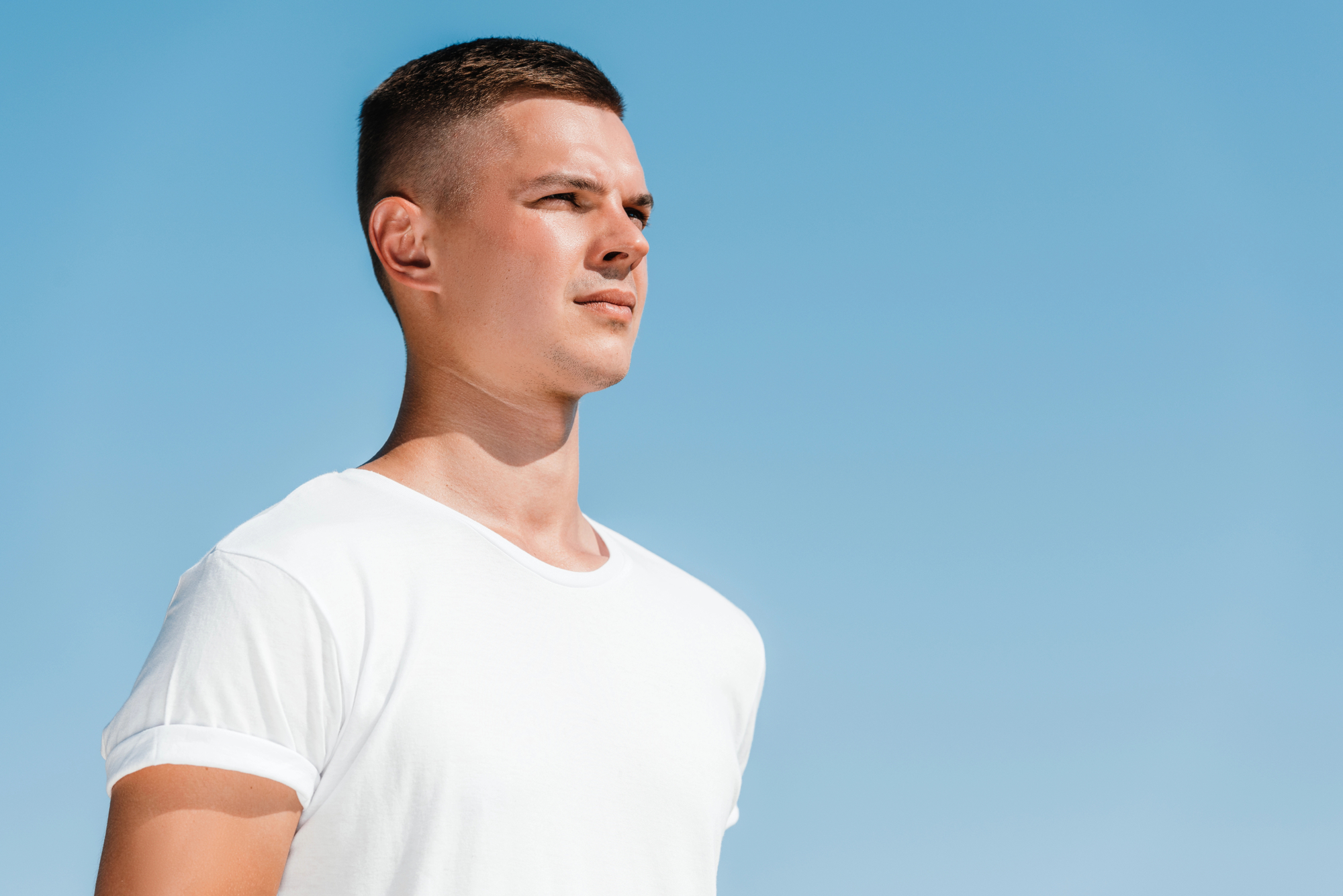 A man with short brown hair wearing a white t-shirt looks into the distance against a clear blue sky.