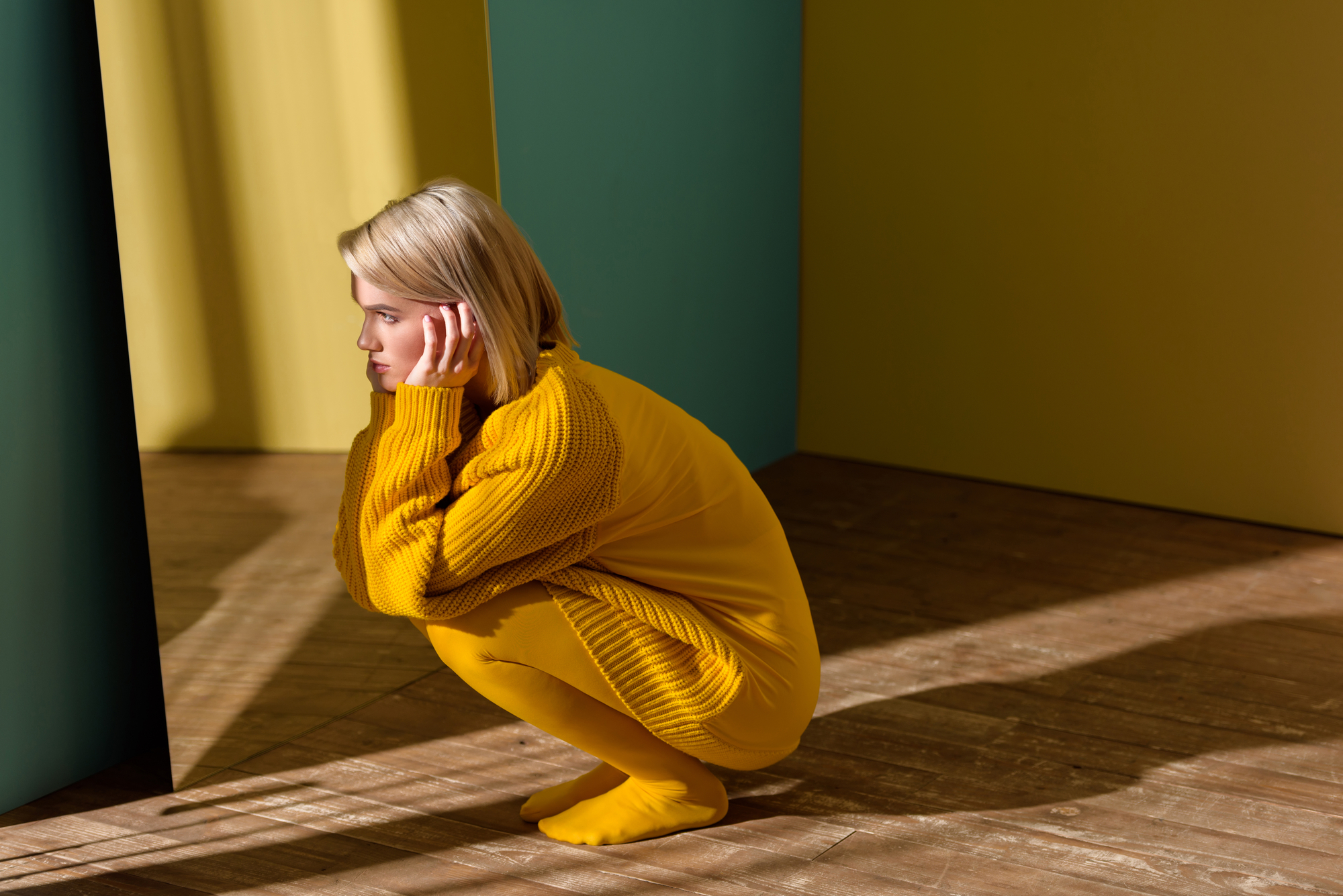 A person with short blonde hair crouches down in a stylish yellow outfit, including an oversized sweater, tights, and socks. They are deep in thought with their head resting on their hand. The background features a mix of green and yellow panels with sunlight casting shadows.