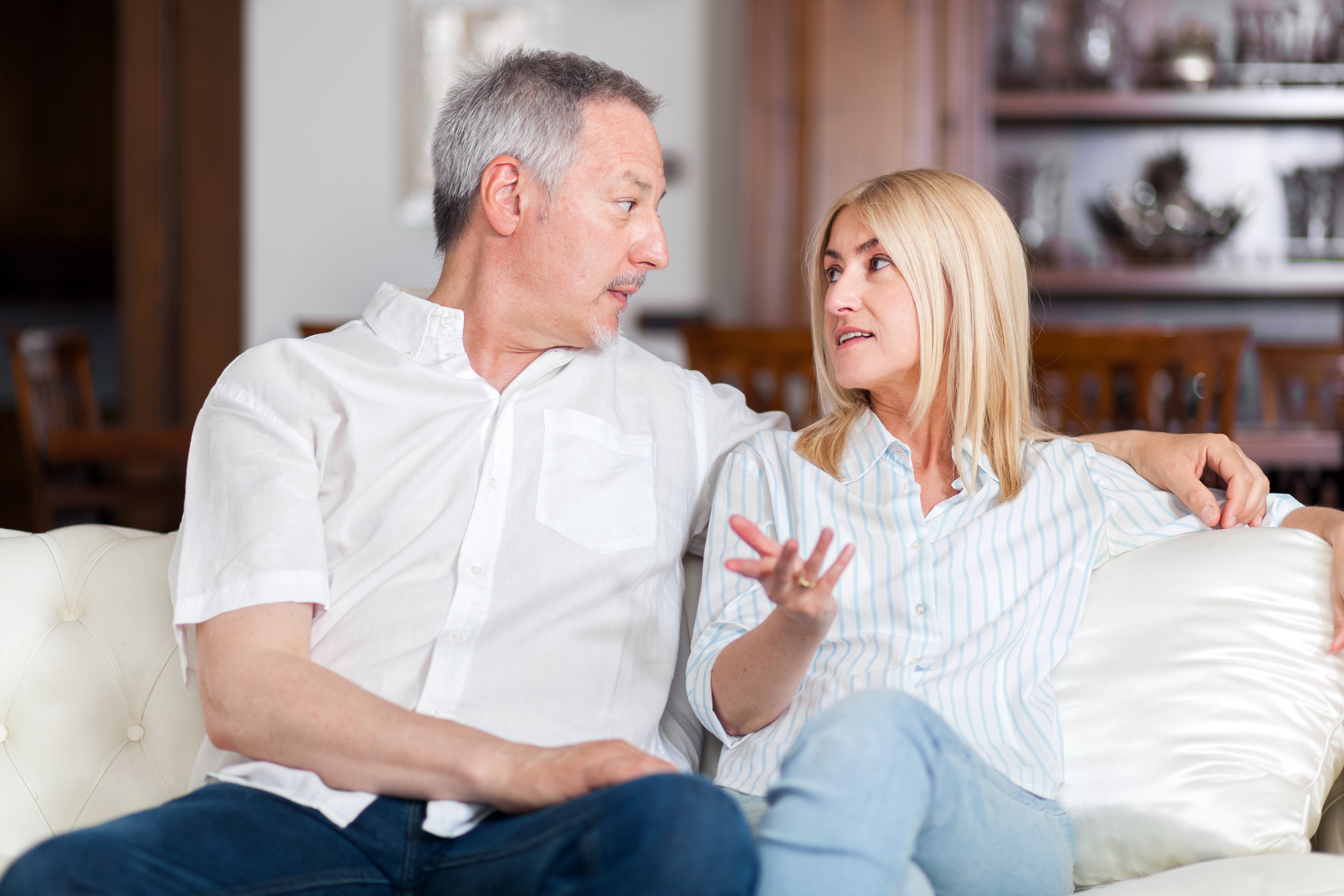A middle-aged couple is sitting on a white couch in a living room. The man, with gray hair, is wearing a white shirt. The woman, with blonde hair, is wearing a light-colored striped shirt. Both are engaged in conversation, with the woman gesturing as she speaks.