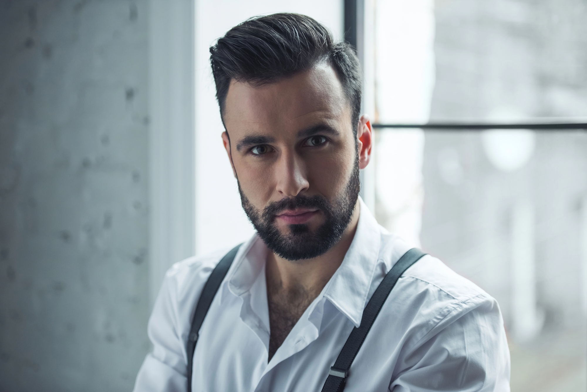 A man with a neatly trimmed beard and styled hair is wearing a white dress shirt with suspenders. He is posing in front of a window, which allows soft, natural light to illuminate his face and creates a serene ambiance in the room with a partially visible brick wall.