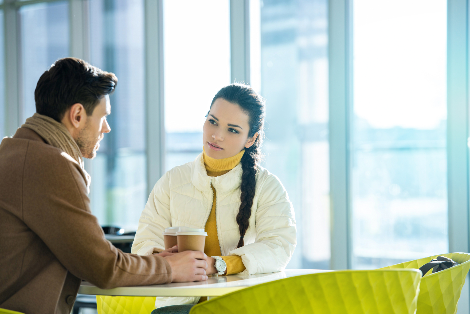 A man and woman sit at a table in a brightly lit cafe, engaged in conversation. The man, wearing a brown coat and scarf, holds a coffee cup. The woman, with a long braid and in a white jacket, listens attentively. Yellow chairs are visible in the foreground.