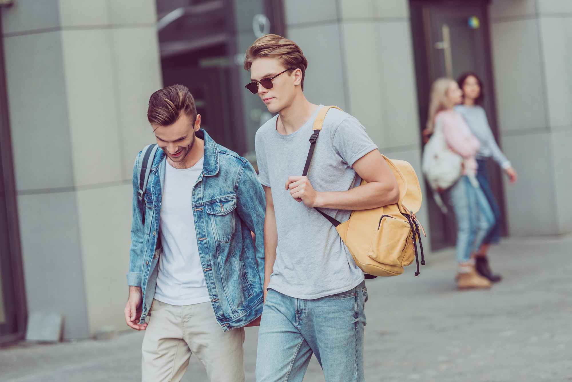 Two young men walking side by side on a city street. The man on the left wears a denim jacket and carries a backpack, while the man on the right wears a gray t-shirt, sunglasses, and a yellow backpack. Two people are conversing in the background.
