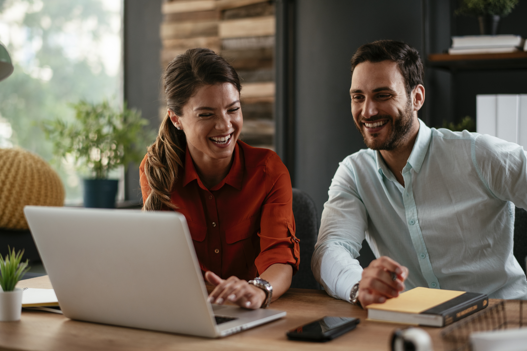 Two people are sitting at a desk and looking at a laptop screen while smiling. The person on the left is wearing a red shirt, and the person on the right is wearing a light blue shirt with a yellow notebook in front of them. They appear engaged and happy.