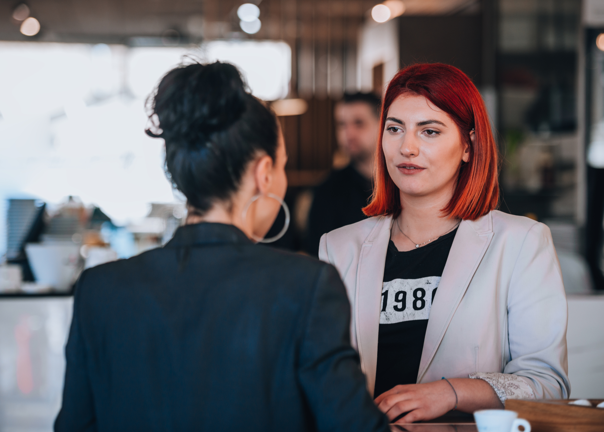 Two women are having a conversation in a cafe. One woman, with red hair and wearing a white blazer over a black shirt with the number "1987," faces the camera. The other woman, with dark hair in a bun, wears a dark jacket and is facing away from the camera.