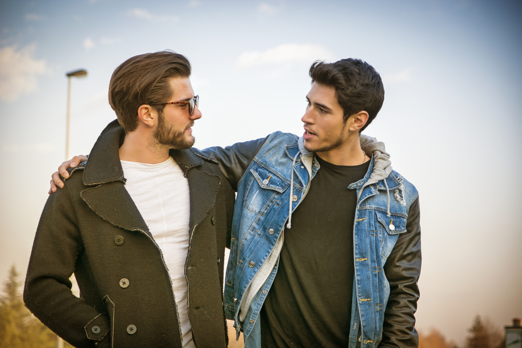 Two young men stand outside against a blue sky with light clouds. One wears sunglasses, a white t-shirt, and a dark coat, while the other wears a denim jacket and a black hoodie. The man in the denim jacket has his arm around the other, and they appear to be talking.