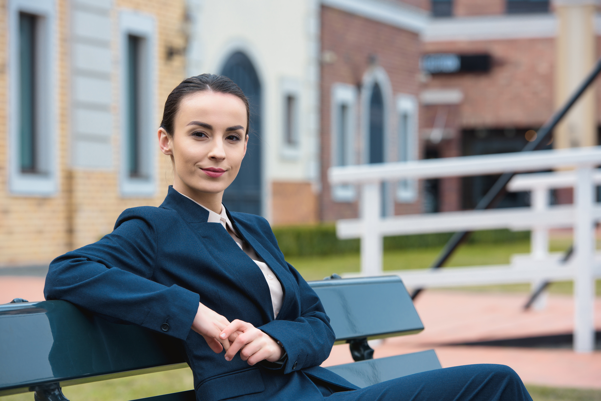 A confident person wearing a navy blue suit is sitting on a bench outdoors. They have short dark hair tied back and are smiling slightly. The background features buildings and a grassy area, suggesting an urban or suburban setting.