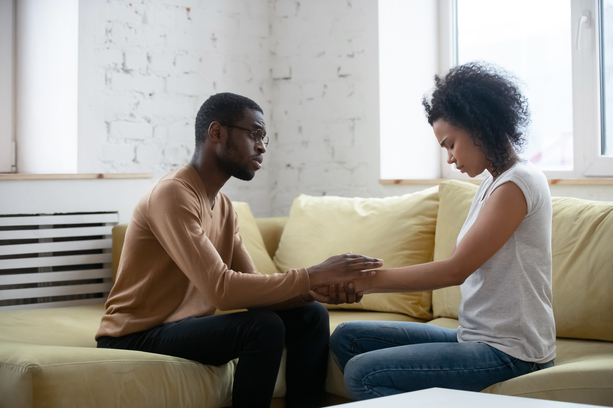 A man and a woman sitting on a yellow sofa in a well-lit room. The man, wearing glasses and a brown sweater, holds the woman's hands while she looks down with a serious expression, wearing a white t-shirt and grey pants. They seem to be having a heartfelt conversation.