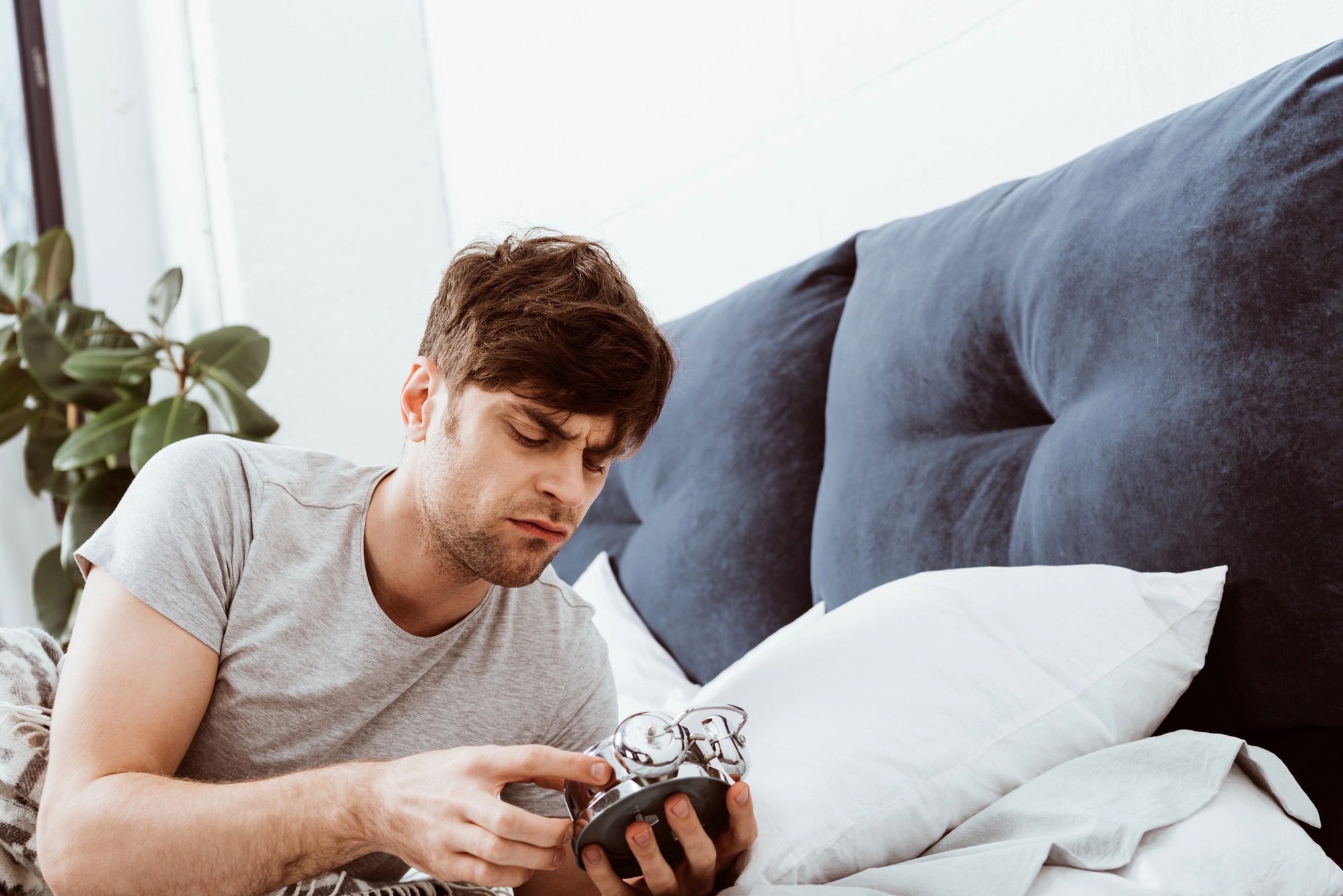 A man lying in bed is looking at an alarm clock with a confused expression. He is holding his glasses with one hand and the clock with the other. There are white pillows and a blue upholstered headboard behind him. A plant is visible in the background.