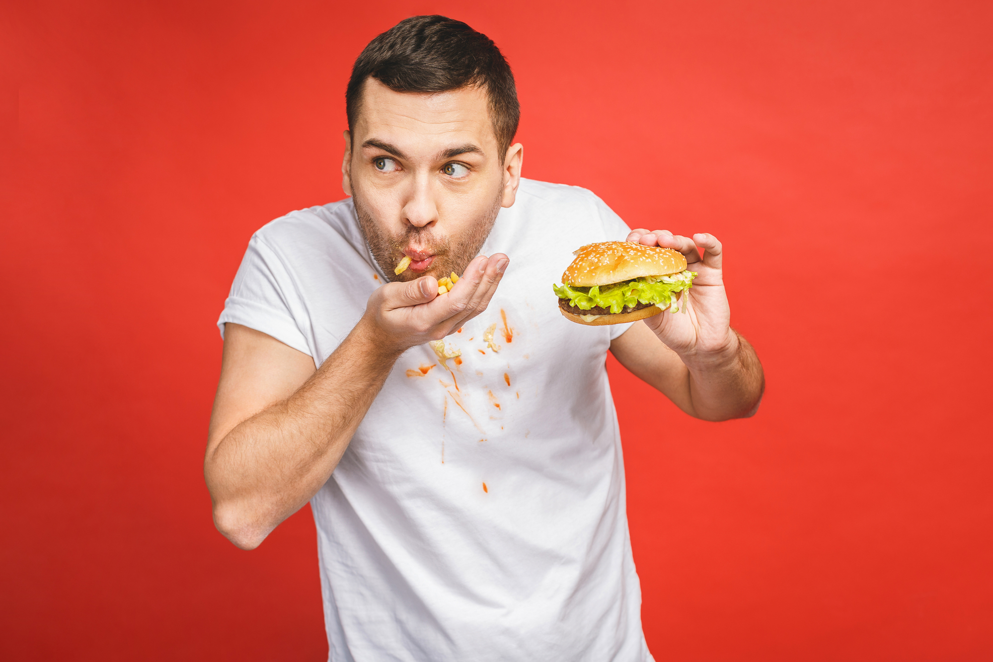 A man in a white t-shirt stands against a bright red background, holding a hamburger in one hand and wiping his mouth with the other. He has food stains on his shirt and an expression of delight while eating.