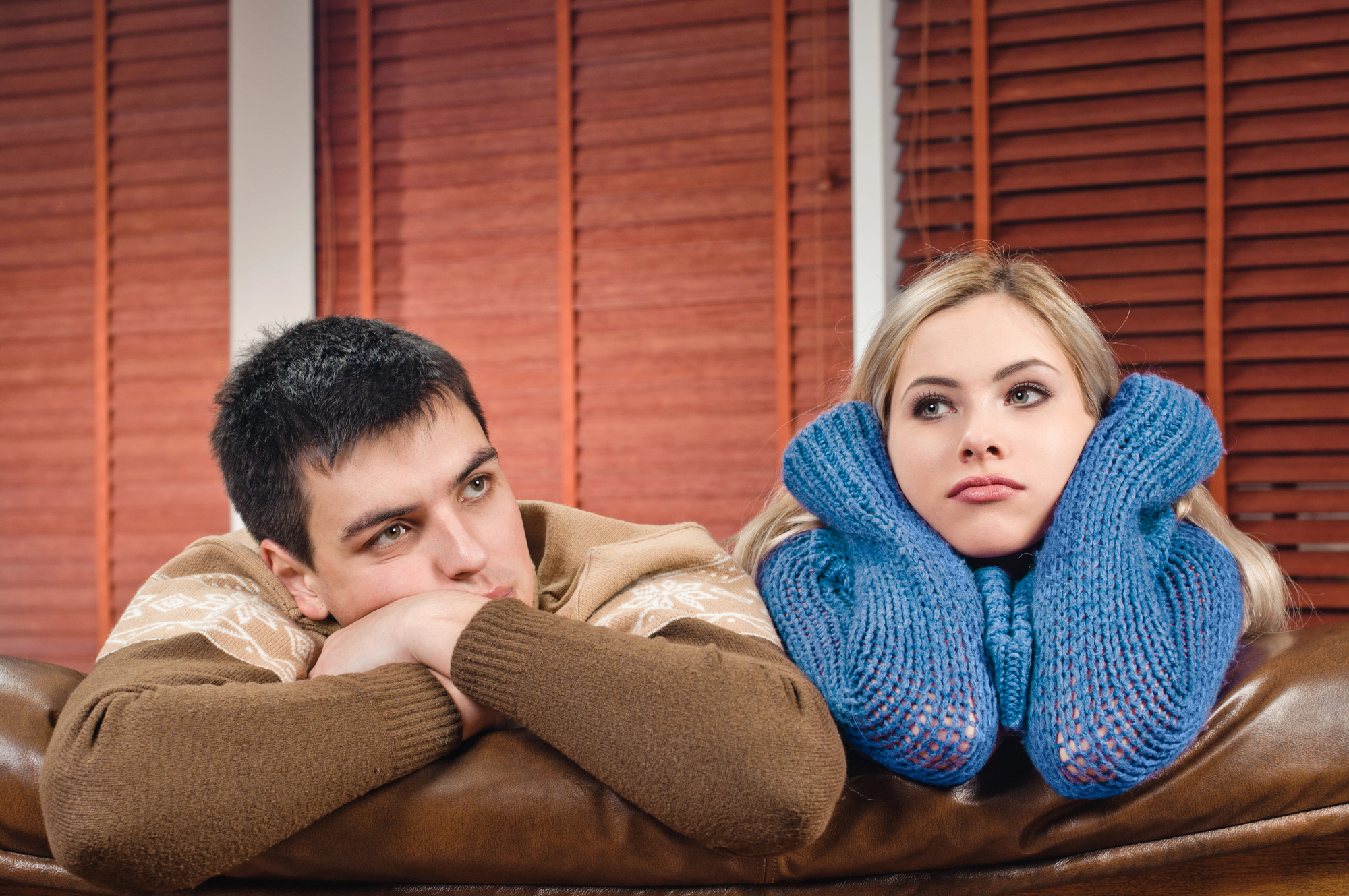 A young man and woman are sitting next to each other on a brown leather couch. The man is wearing a brown sweater and has his chin resting on his arms, looking pensively ahead. The woman, wearing a blue knit sweater, has her head resting on her gloved hands, appearing contemplative. Brown blinds cover the windows behind them.