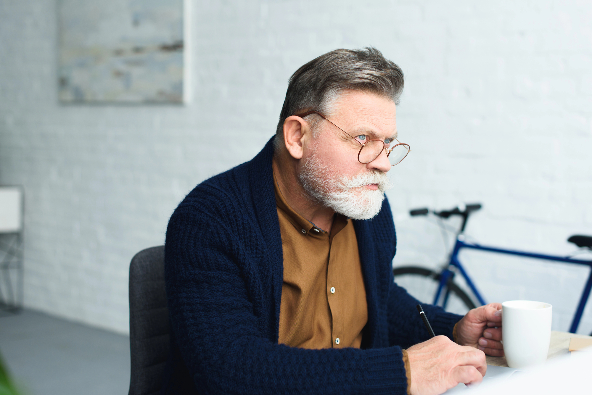 An elderly man with glasses and a beard, wearing a brown shirt and blue cardigan, sits at a desk holding a white coffee mug in one hand and writing with the other. A bicycle and a blurred painting are visible in the background.