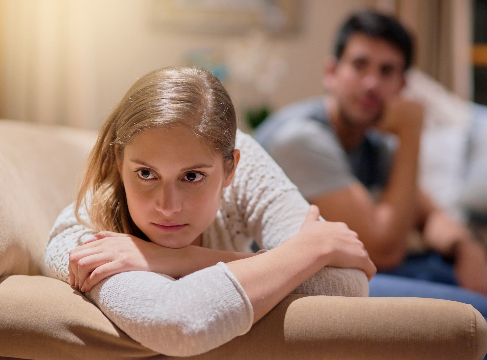 A woman with blonde hair looks upset and rests her head on her crossed arms on the back of a couch. In the blurred background, a man looks in her direction, appearing concerned. The room is softly lit, creating a warm ambiance.