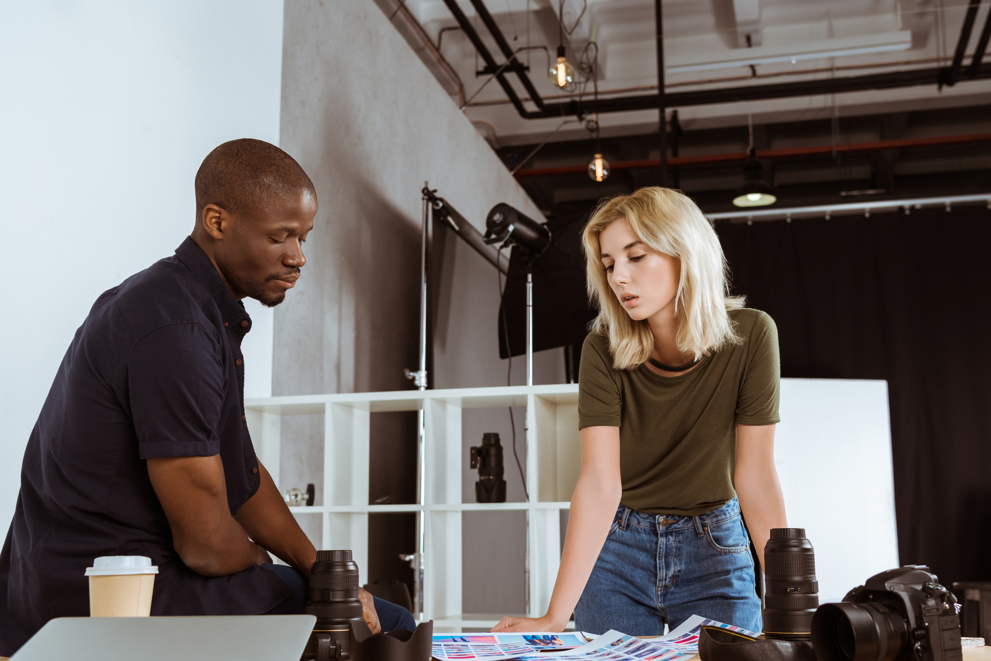 Two people, a man and a woman, are intently reviewing photographs on a table in a modern studio. Camera equipment, including lenses and a camera, is scattered on the table. The background features shelves, lighting equipment, and a minimalistic design.