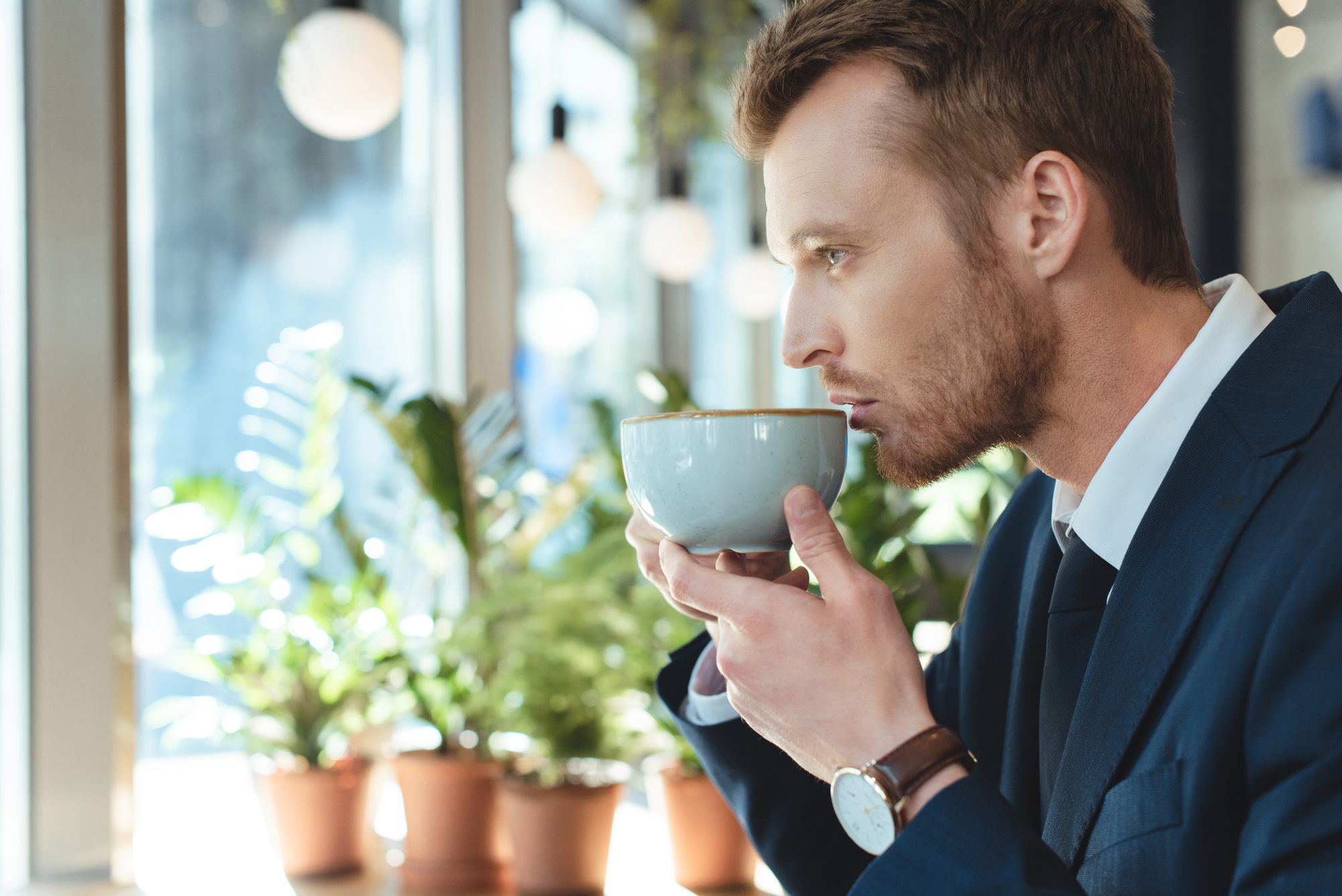 A man wearing a dark suit sips from a light-colored cup while sitting by a window. Sunlight filters through the glass, illuminating potted plants on the windowsill. The man looks thoughtful as he enjoys his drink.