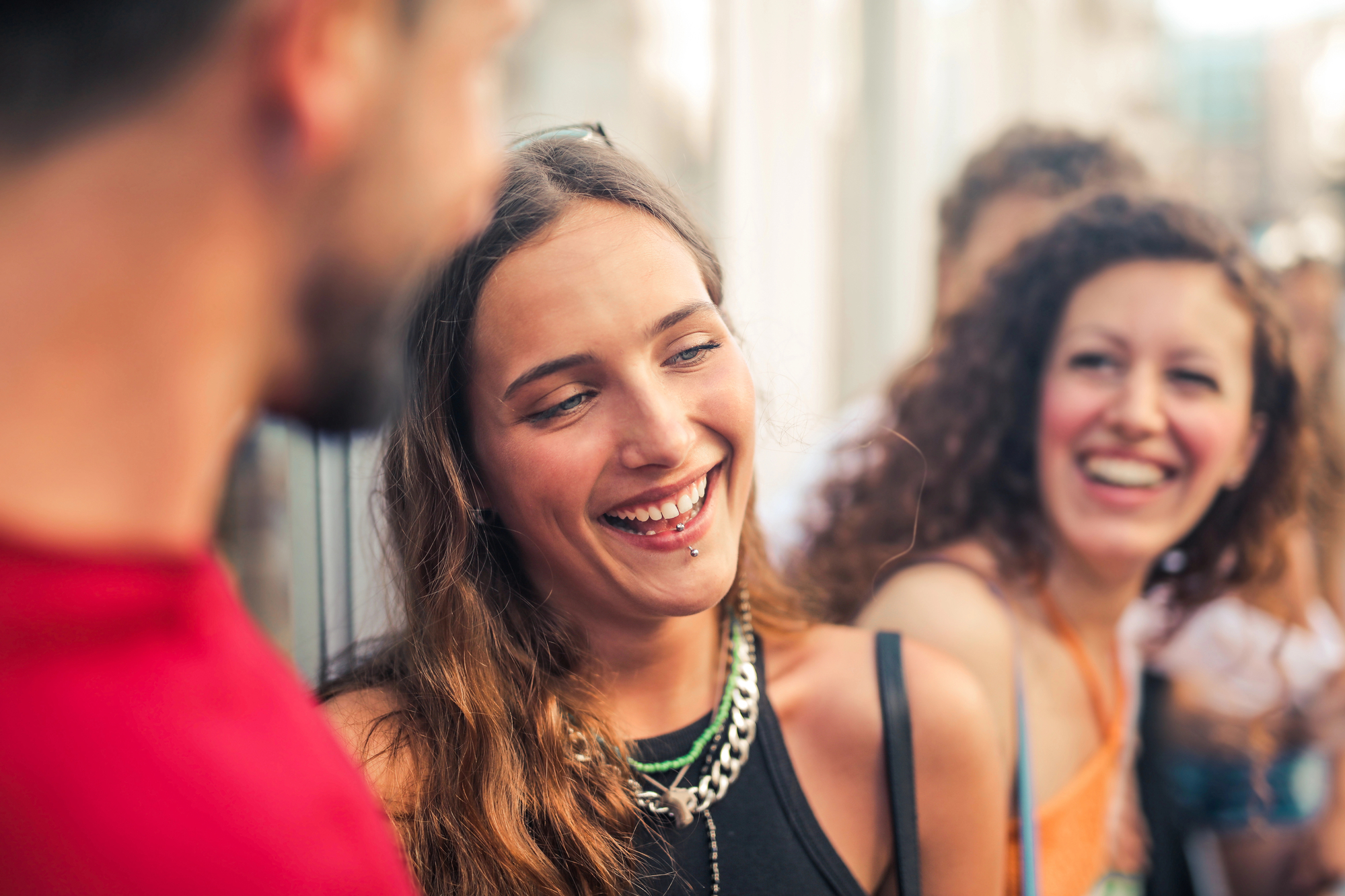 A group of young adults socializing and laughing together outdoors. One woman with long hair and wearing a black top and silver necklace is in focus, smiling warmly towards the person next to her, while another woman with curly hair laughs in the background.