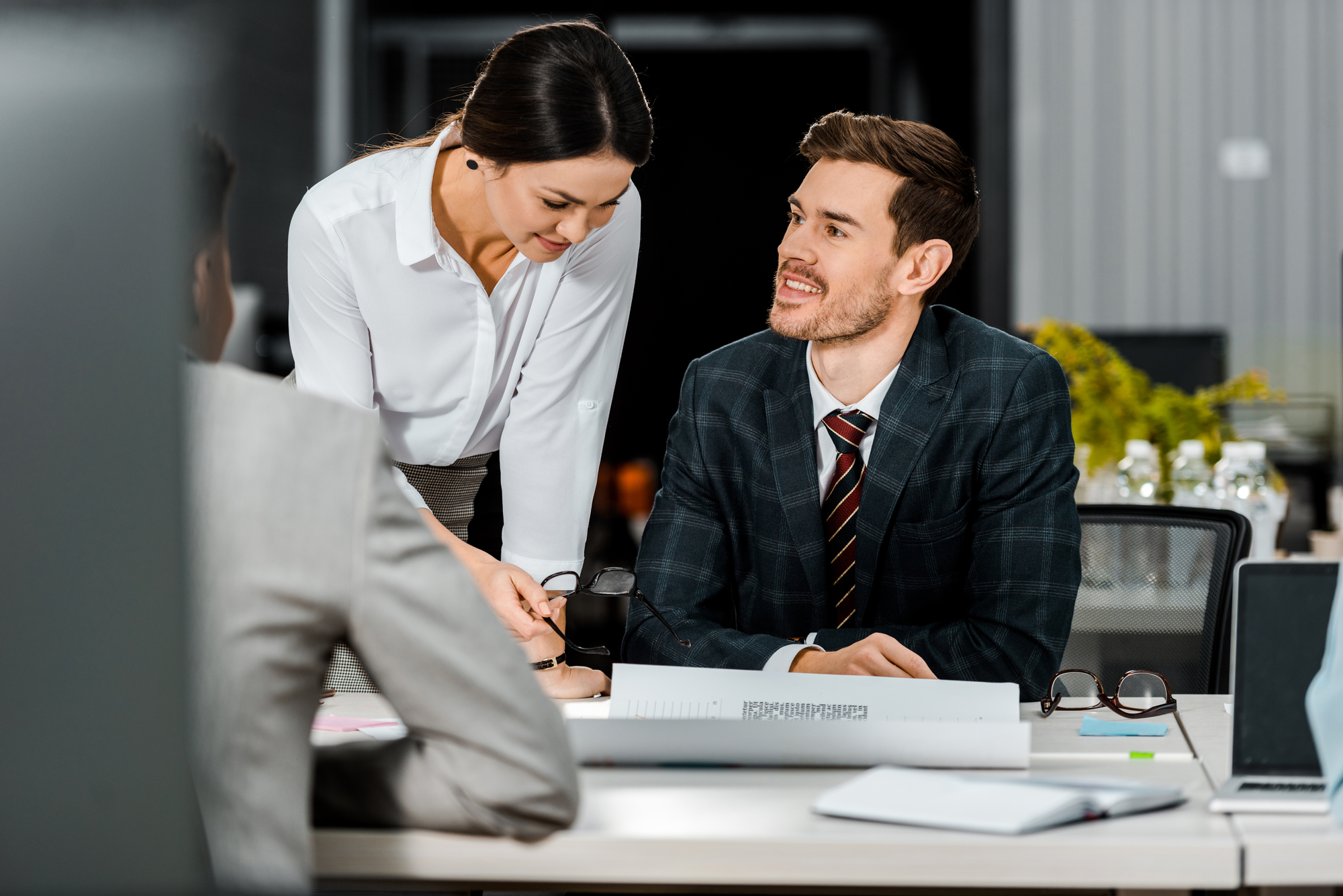 A man in a suit smiles and looks at a woman in a white blouse who is leaning over the table, pointing at a document. They are having a discussion in a modern office with a blurred background, including another person out of focus in the foreground.