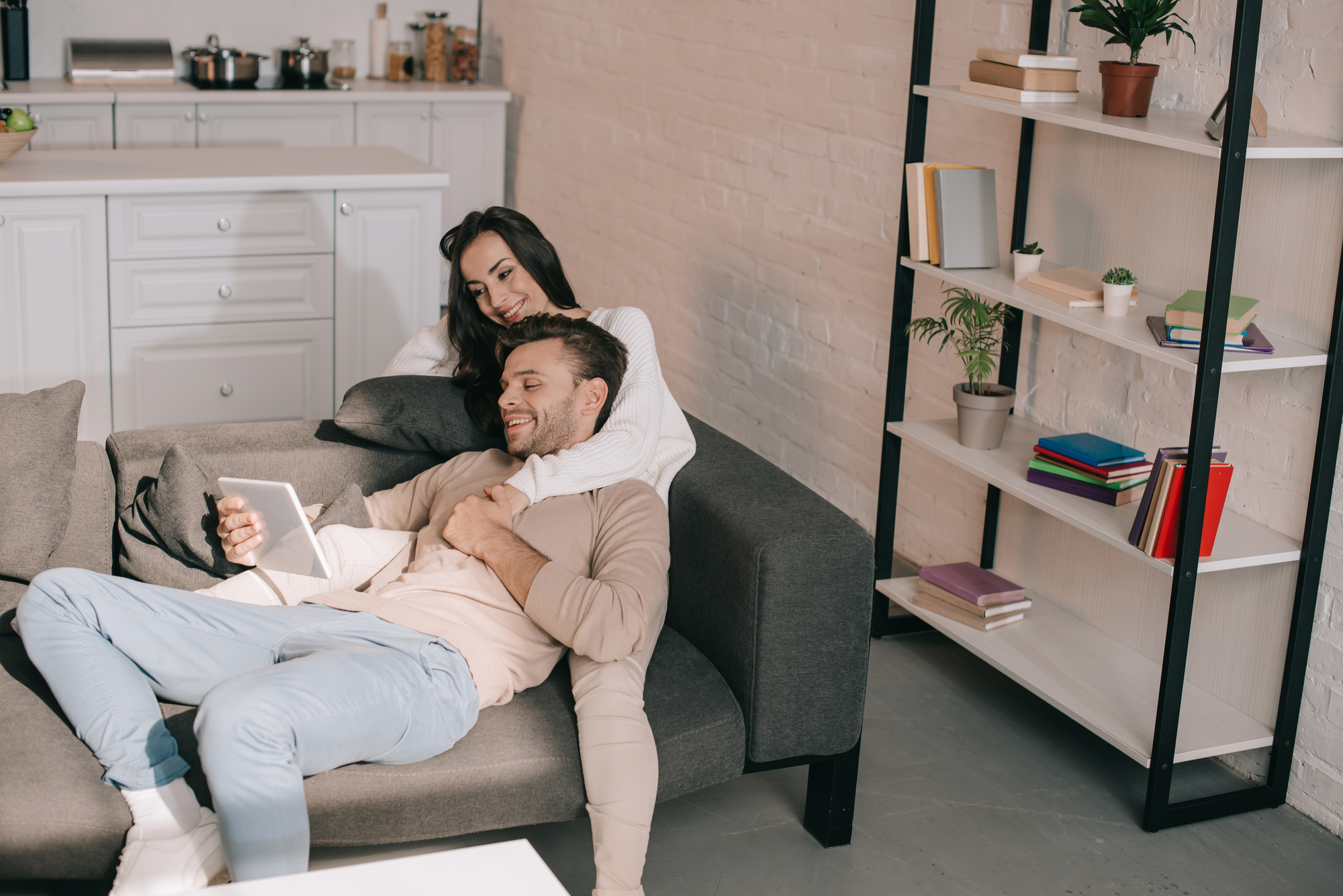 A couple relaxes on a gray sofa in a modern living room. The woman, with long dark hair, sits upright and embraces the man, who is lying down and holding a tablet. Behind them, a white kitchen and a white shelf with colorful books and potted plants are visible.