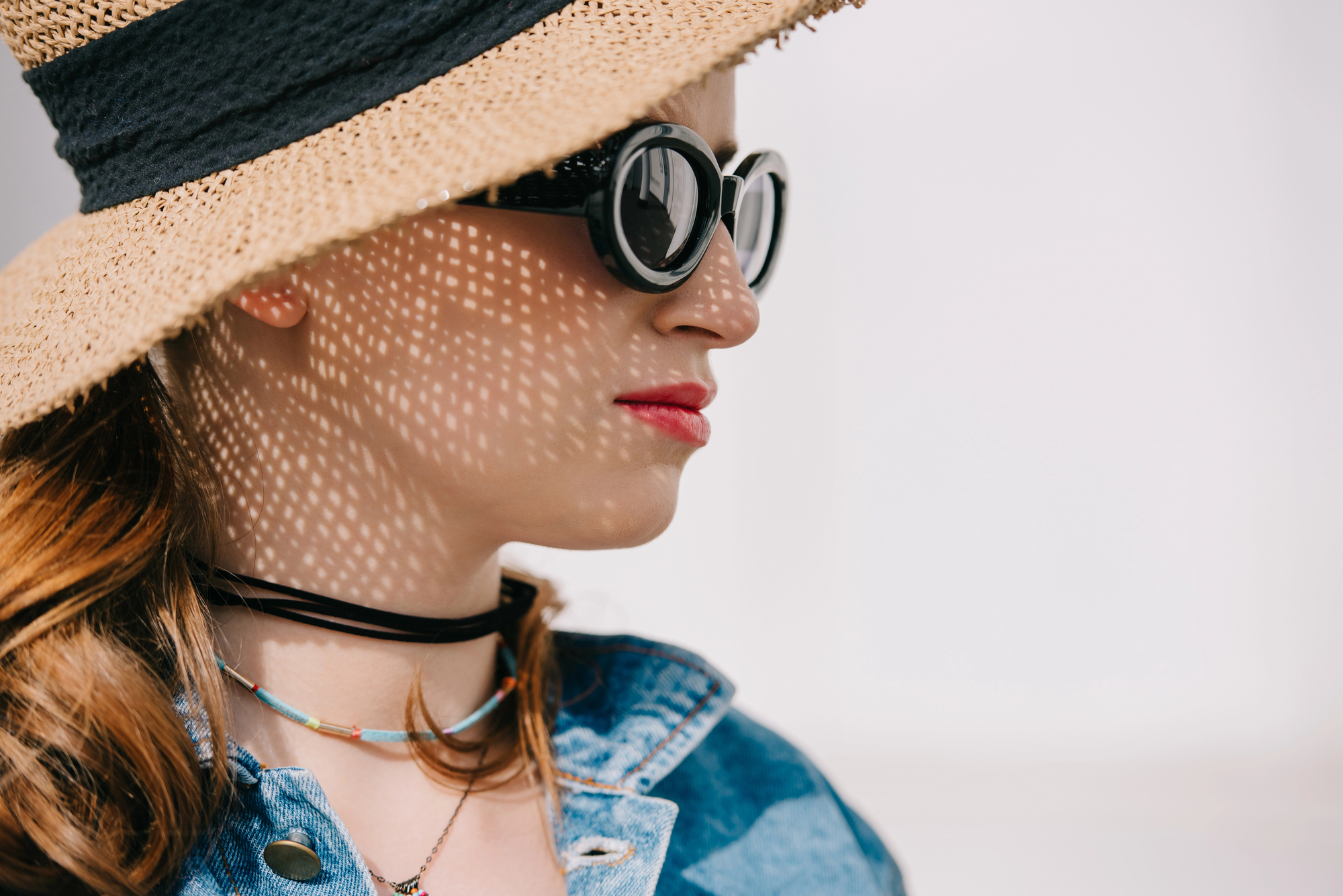 A person wearing a wide-brimmed straw hat and round black sunglasses stands against a bright background. The hat casts intricate shadow patterns on their face. They wear a denim jacket, a multi-layered necklace, and have long, loose hair.