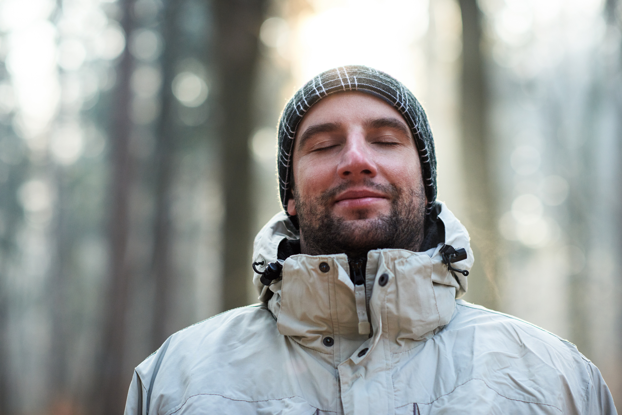 A person stands in a forest, eyes closed, with a peaceful expression. They are wearing a warm, light-colored jacket and a dark beanie with a checkered pattern. Sunlight filters through the trees in the background.