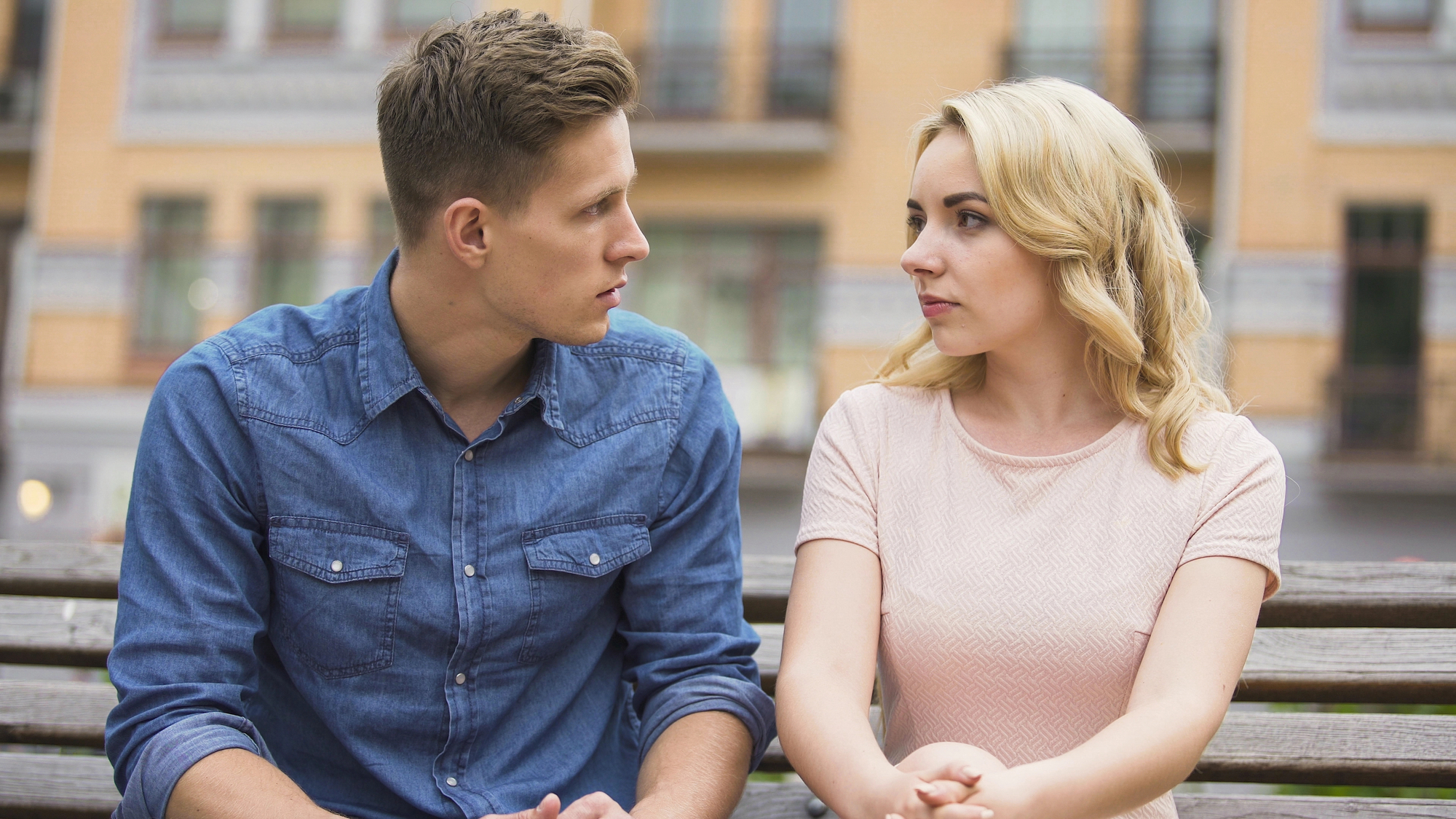 A young man wearing a blue denim shirt and a blonde woman in a light pink top sit close together on a bench outdoors. They are looking intently at each other, appearing engaged in a serious conversation. The background features blurred buildings.