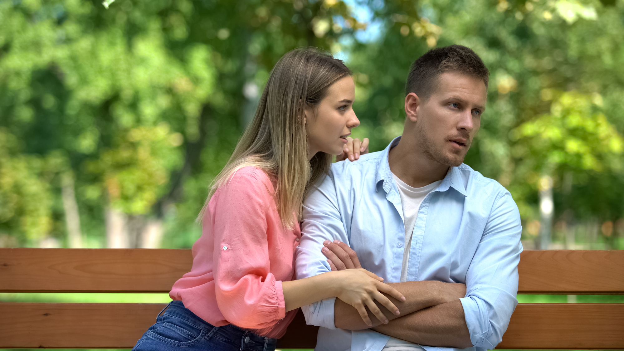 A woman with a concerned expression leans towards a man with folded arms, both seated on a park bench. She appears to be talking or comforting him, while he looks away thoughtfully. Green trees and foliage are visible in the blurred background.