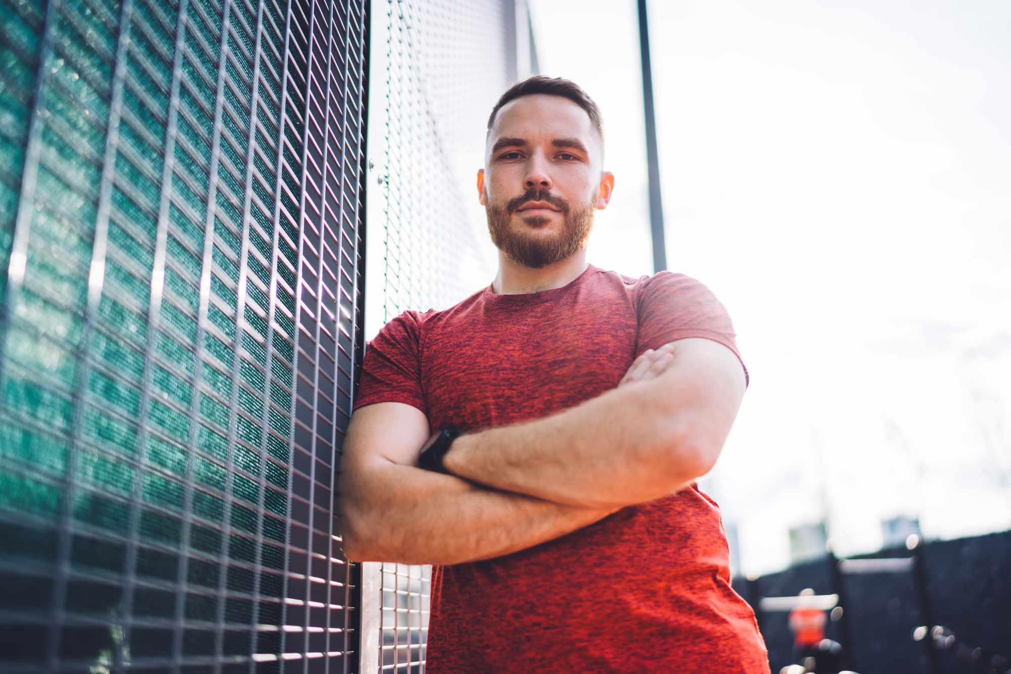 A man with a trimmed beard and short hair stands with his arms crossed, leaning against a metal fence. He wears a red t-shirt and looks directly at the camera with a confident expression. Bright sunlight creates a vivid backdrop, with a slightly blurred urban environment.