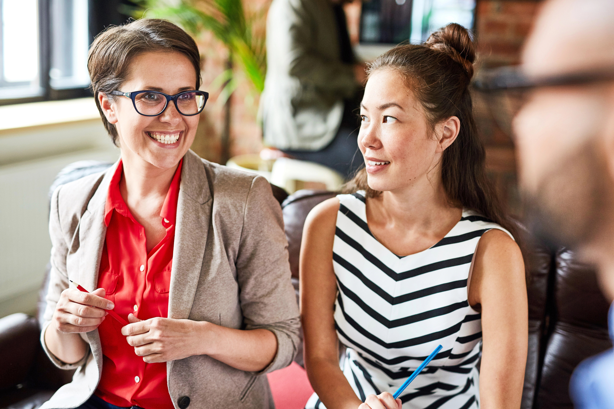 Two women are seated and smiling in an office setting. The woman on the left wearing glasses and a beige blazer over a red top, holds a pen. The woman on the right has long hair tied back and is dressed in a black-and-white striped dress, also holding a pen.