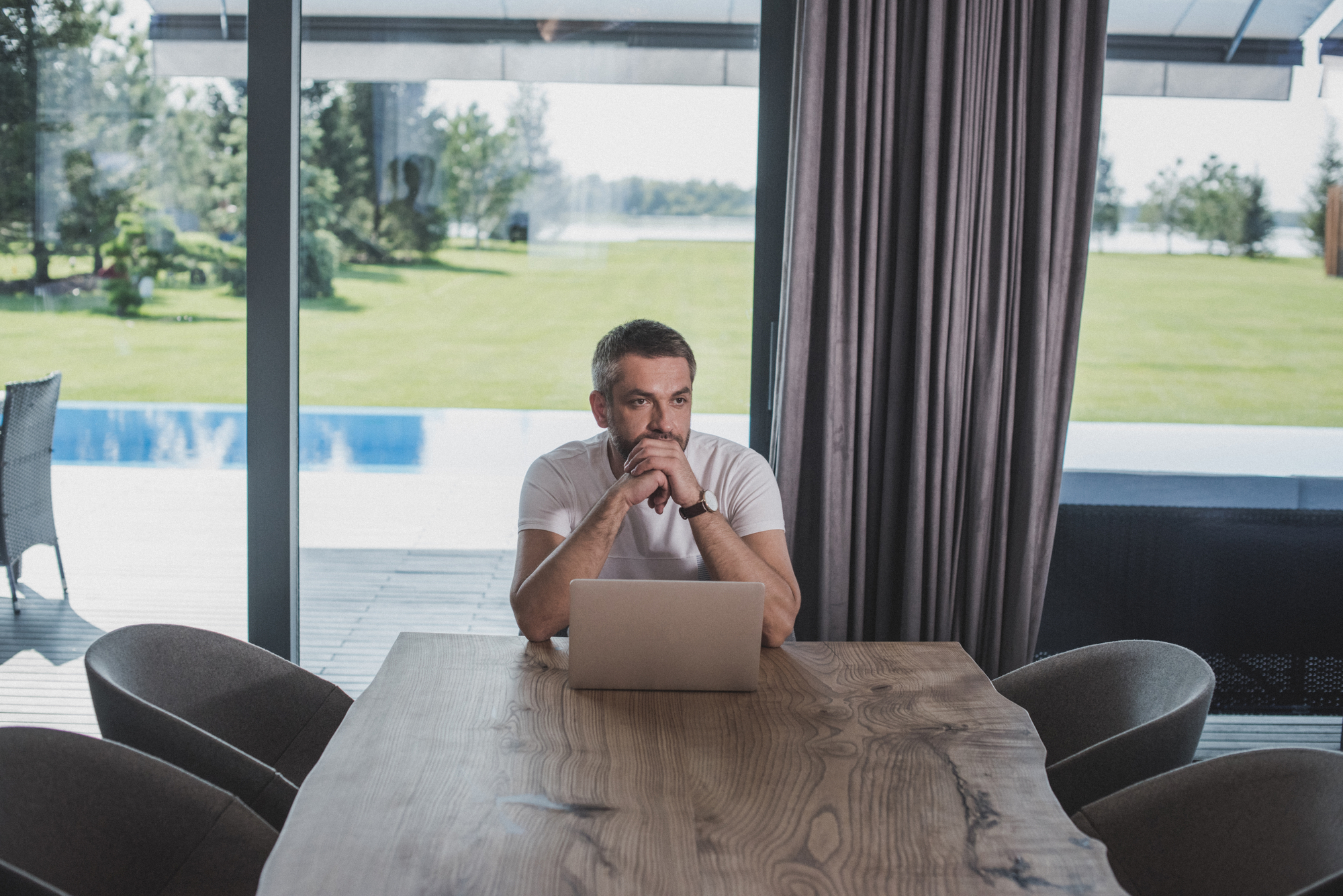 A man sits at a wooden table in front of a laptop in a bright room with large windows. He rests his chin on his clasped hands, looking thoughtfully into the distance. The view outside includes a lush green lawn and a pool, partially obscured by curtains.