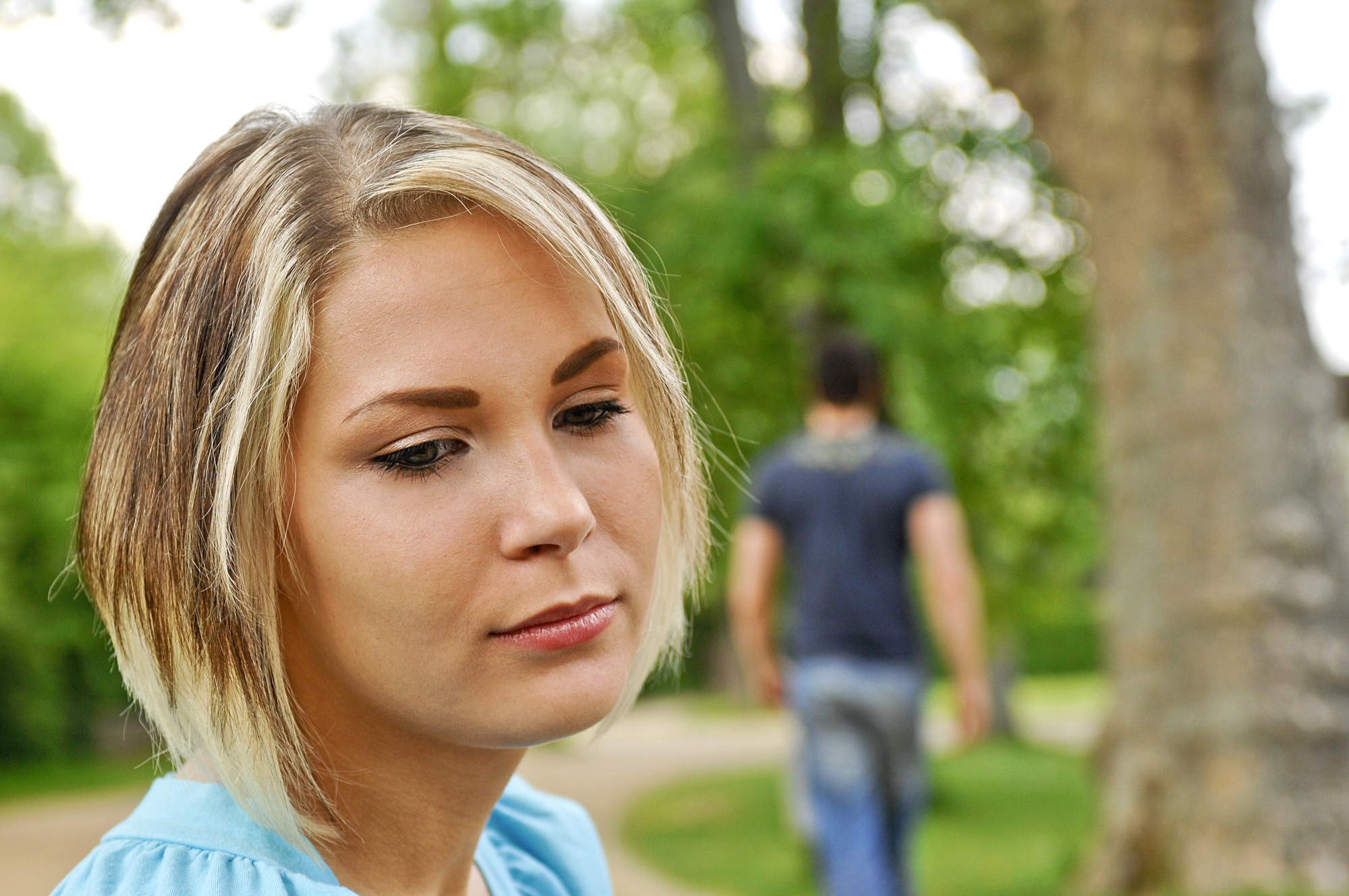 A woman with short blonde hair and a blue shirt appears sad while sitting outdoors. In the background, a man wearing a dark shirt and jeans walks away on a path through a green, leafy park.