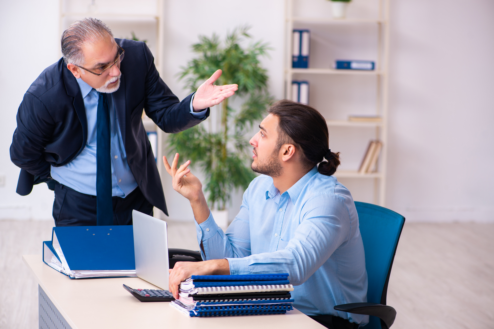 An older man in a suit is gesturing and talking to a younger man with a beard and ponytail, who is sitting at a desk with an open laptop, stacks of notebooks, and a calculator. The younger man is listening attentively, holding a pen in one hand.