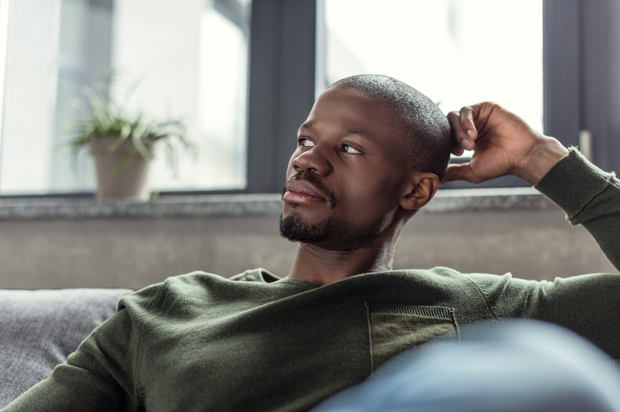 A person in a green sweater is sitting indoors, resting their head on their hand while gazing to the side with a thoughtful expression. A small potted plant is visible on a windowsill in the background.