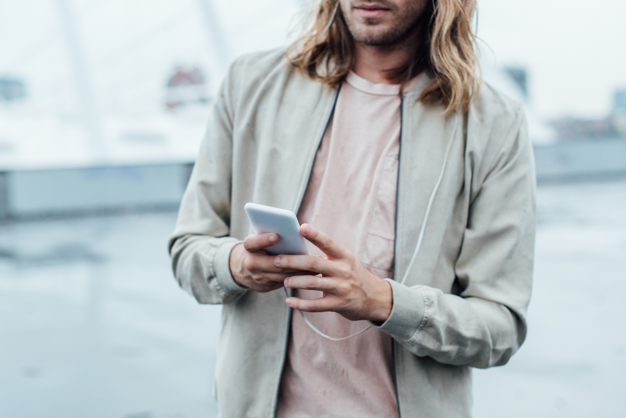 A person with long hair, wearing a light jacket and a pale pink shirt, is holding a white smartphone and appears to be texting or scrolling. They have earphones plugged into the phone, and the background is blurred, showing an outdoor setting.