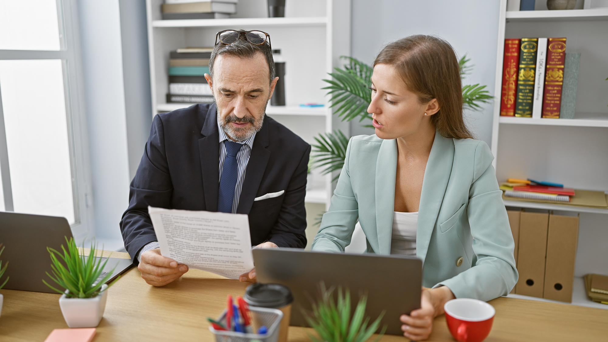 Two people are working together in an office. The man, in a suit and glasses, is holding a document and discussing it with the woman, who is using a laptop. They are surrounded by office supplies, plants, bookshelves, and a window in the background.