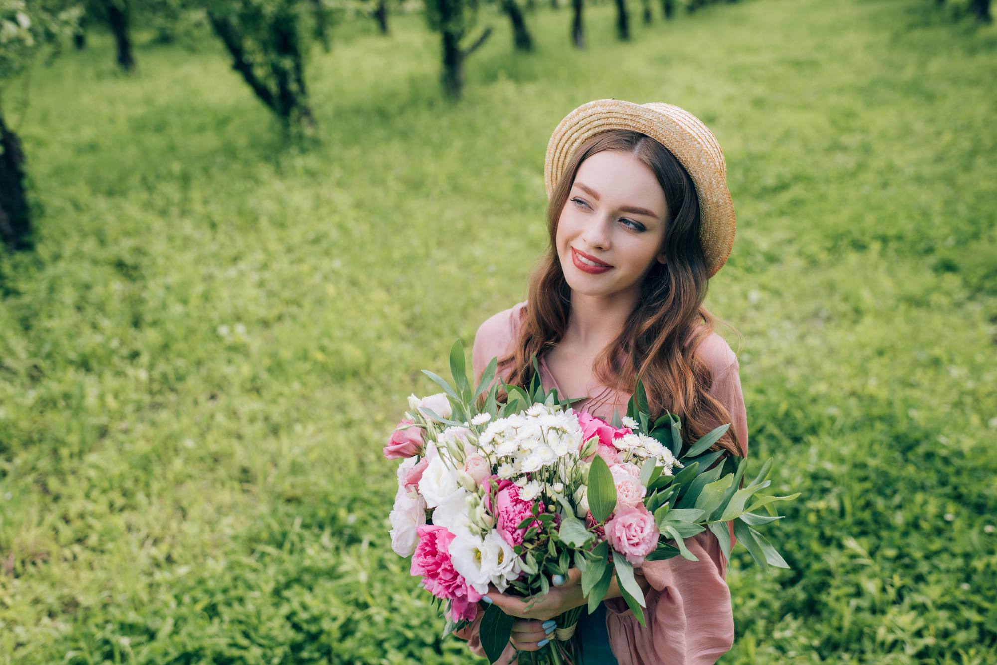 A young woman with long hair wearing a straw hat holds a bouquet of flowers and smiles while standing in a lush green field. She wears a pink top and appears to be enjoying the outdoors, surrounded by greenery.