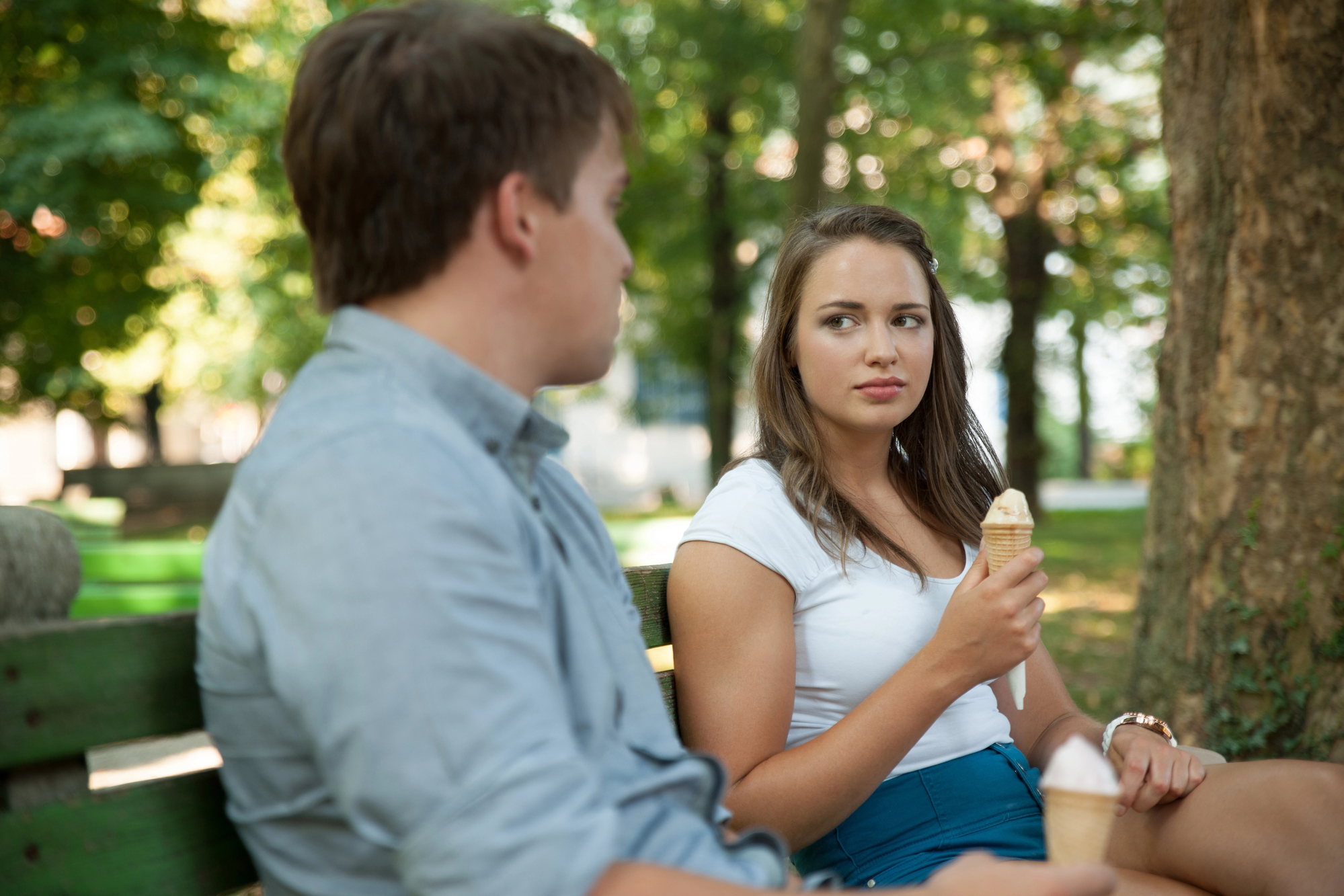 A young man and woman are sitting on a bench in a park, each holding an ice cream cone. The woman, wearing a white shirt and blue skirt, looks at the man with a serious expression. The man, in a light gray shirt, appears to be speaking to her. Trees are in the background.