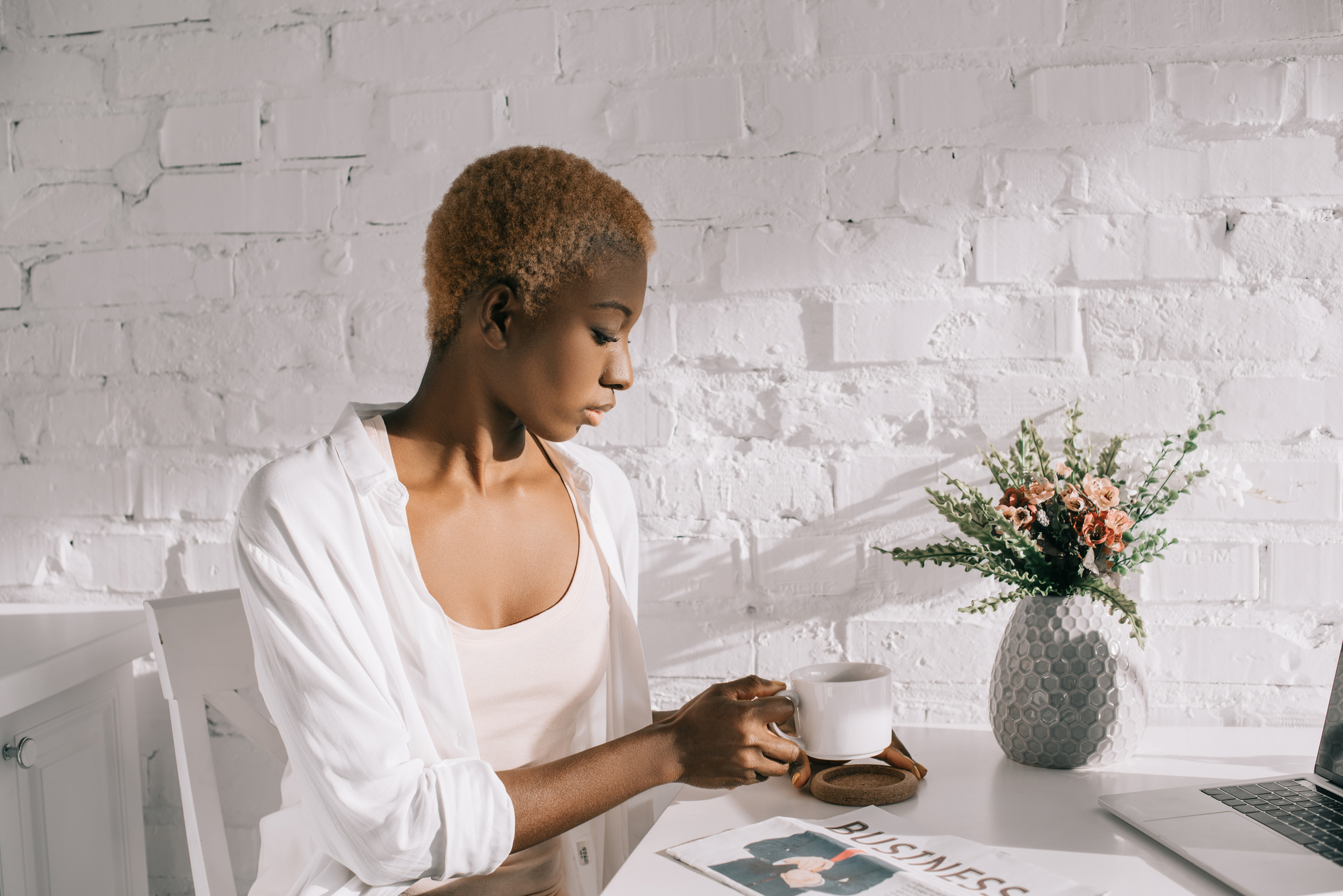A person with short hair sits at a white desk, holding a cup and looking down. They are dressed in a white shirt and beige top. On the desk, there is a magazine titled "BUSINESS," a laptop, and a vase with flowers. The background features a white brick wall.