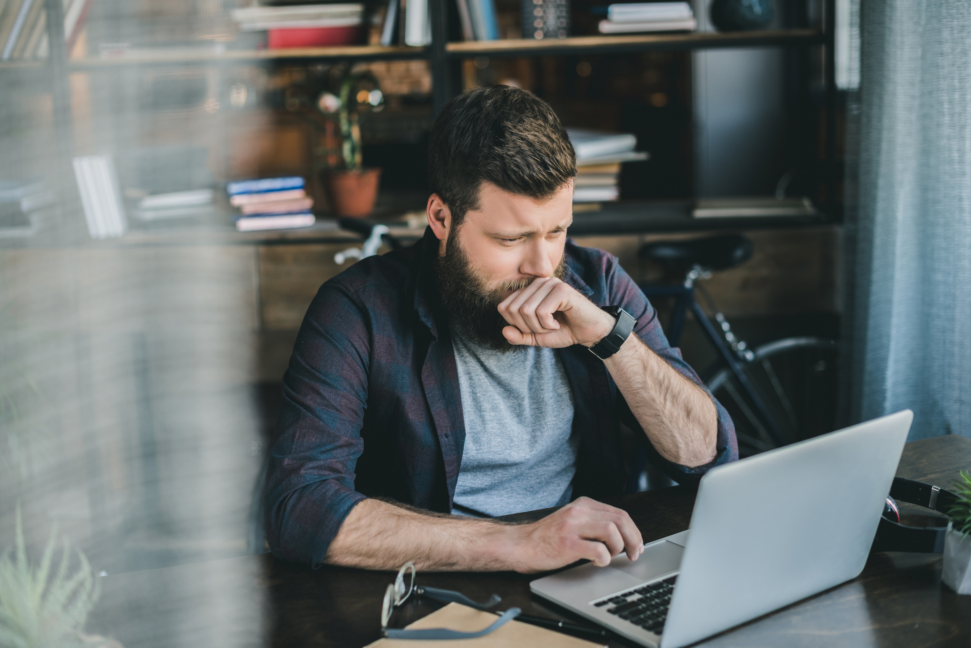 A bearded man sits at a desk in front of a laptop, covering his mouth with his hand as if yawning or coughing. He is dressed casually in a plaid shirt and a gray t-shirt, with a bicycle and bookshelf in the background. The scene suggests a home office or study environment.
