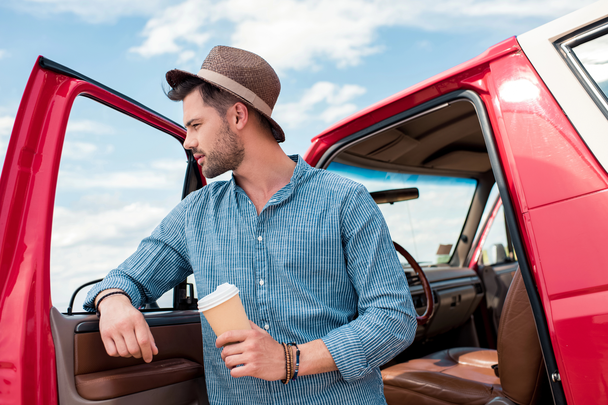 A man in a straw hat and blue checkered shirt leans against the open door of a red car, holding a to-go coffee cup. He is looking to his right with a thoughtful expression. The sky is clear with scattered clouds in the background.