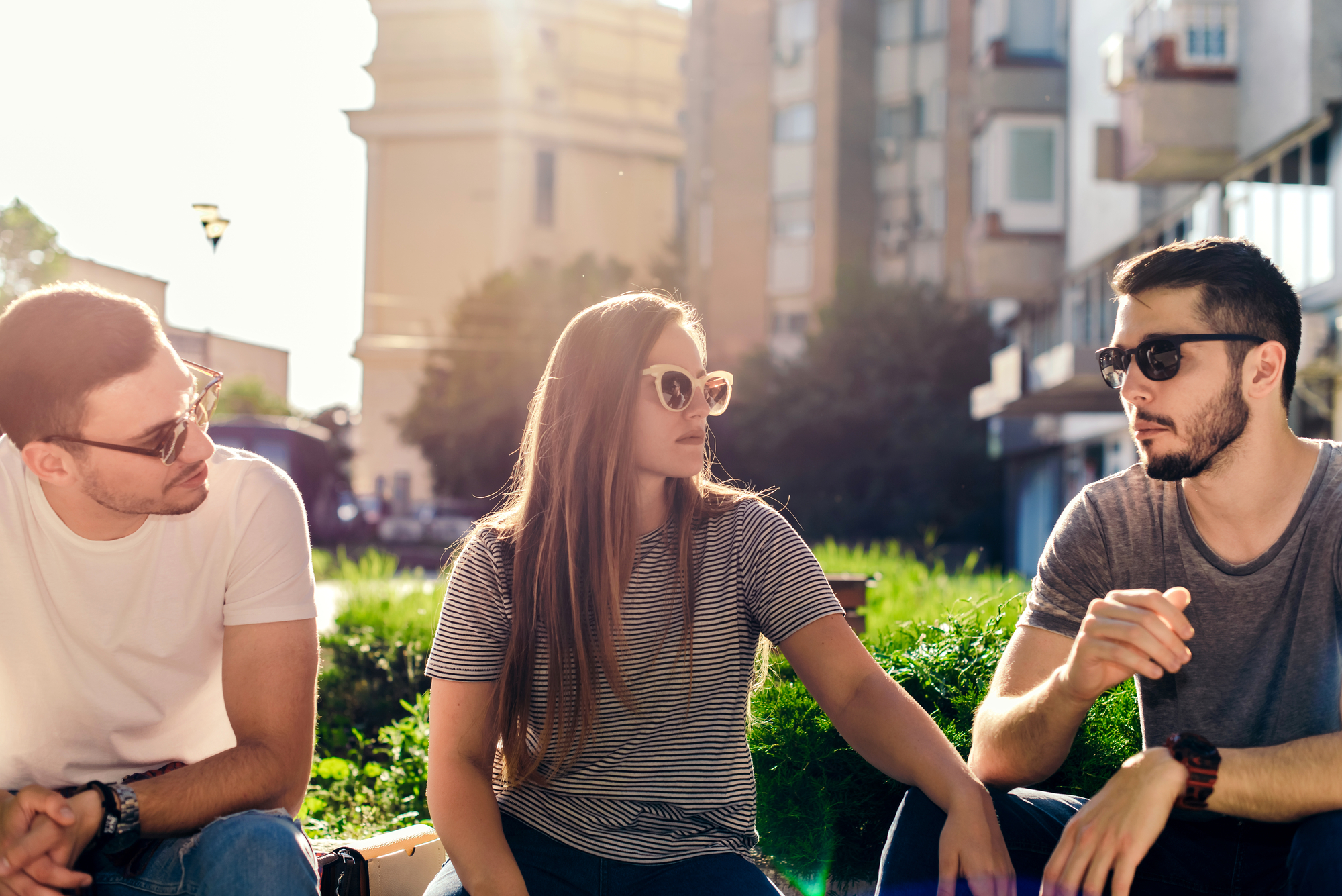Three people sitting on an outdoor bench in the sunlight. The two men, both wearing sunglasses, face a woman in the middle, who is also wearing sunglasses. They appear to be engaged in conversation, with a blurred urban background visible behind them.
