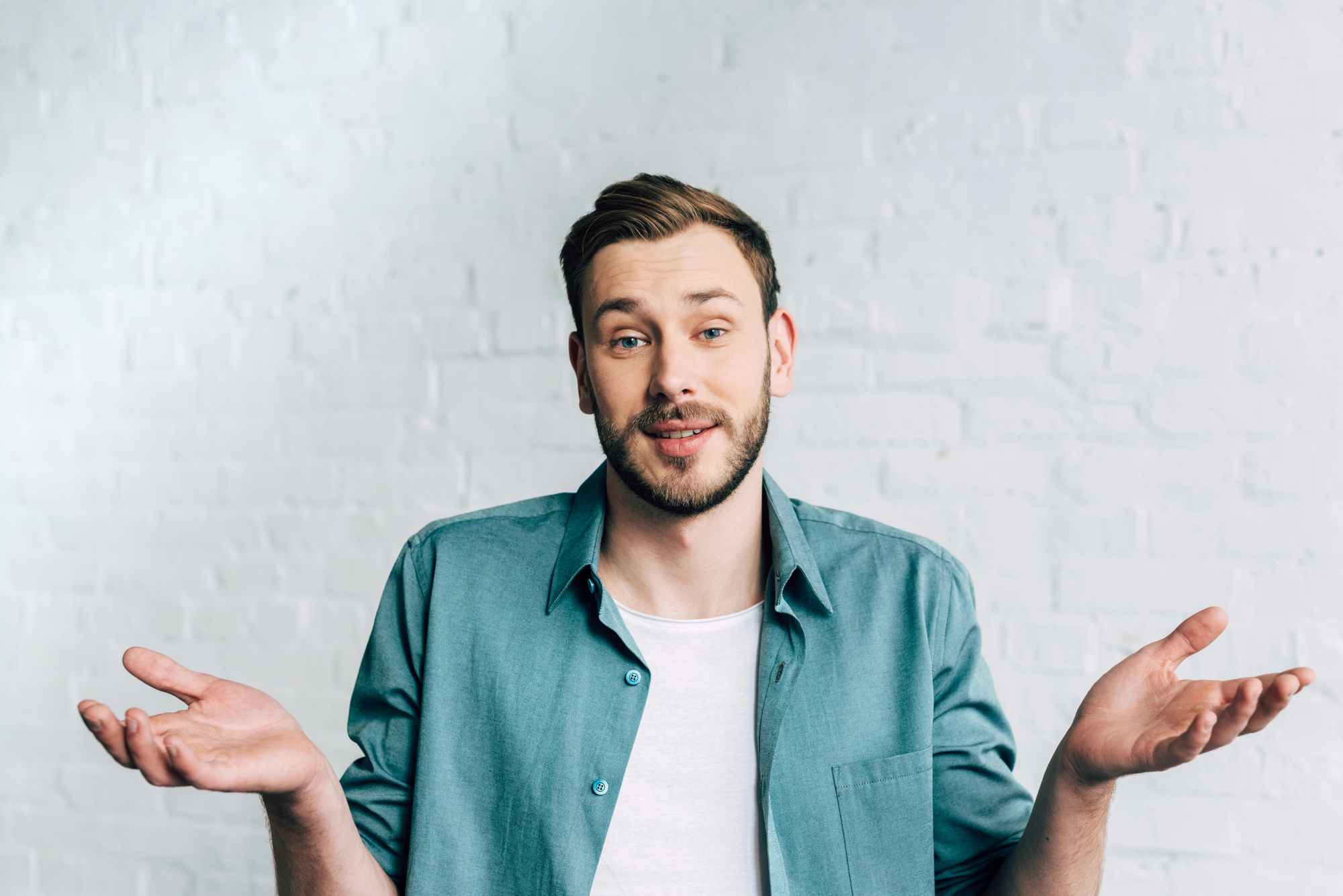 A man with short brown hair and a beard is standing in front of a white brick wall. He is wearing a teal button-up shirt over a white t-shirt and has a slightly amused expression while shrugging with both hands up, palms facing upwards.