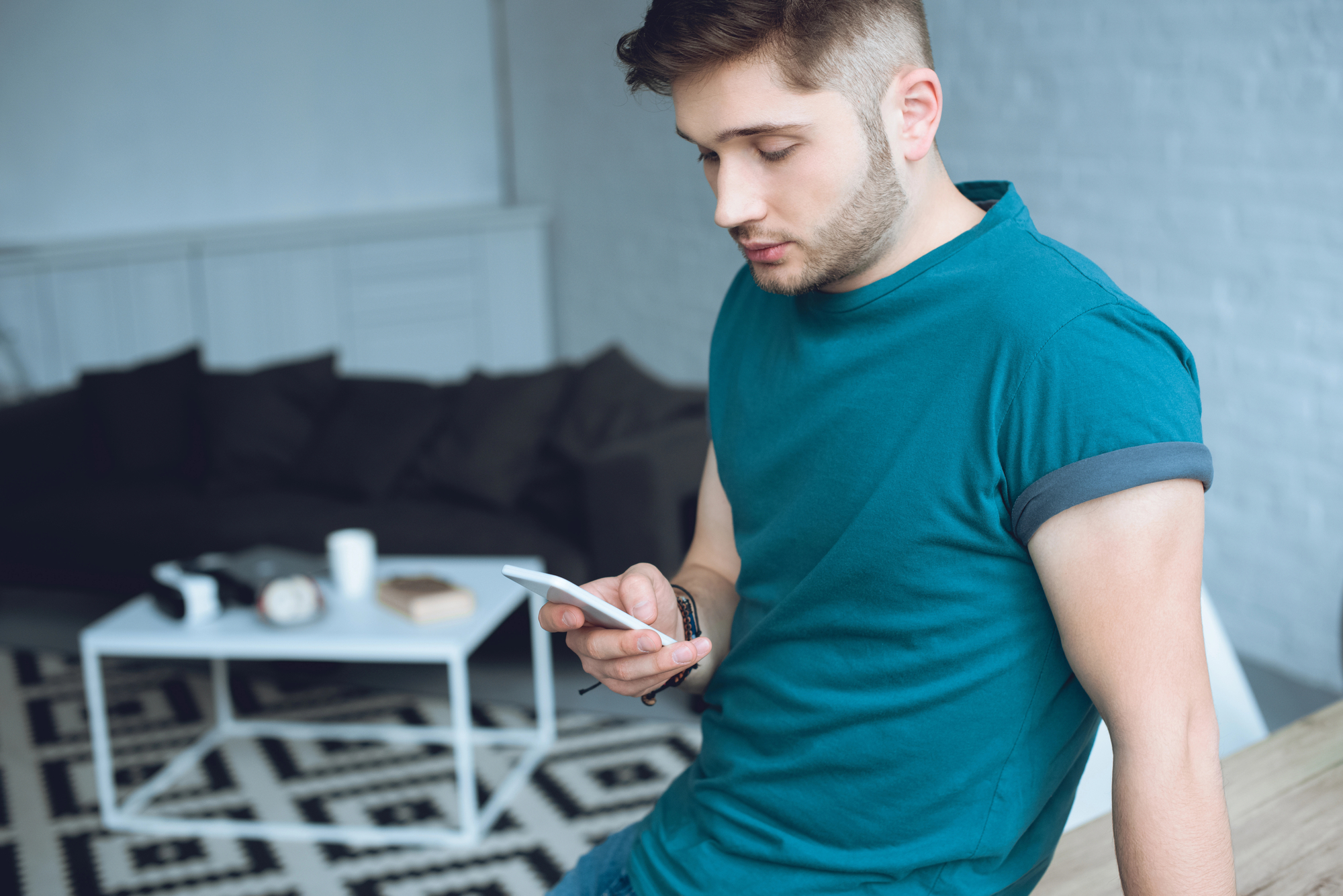 A young man with short hair and a trimmed beard wearing a teal t-shirt is leaning against a table while looking at his smartphone. He is in a modern living room with a white coffee table, black couch, and a geometric-patterned rug in the background.