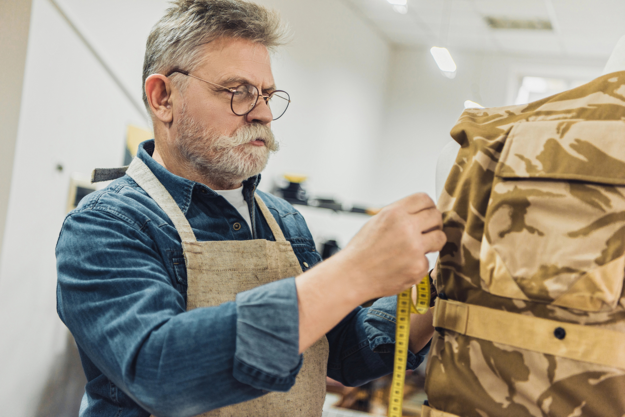 An elderly man with glasses and a mustache wearing a denim shirt and a beige apron is measuring a camouflage jacket using a yellow measuring tape. He appears to be in a workshop or studio with various tools and materials in the background.