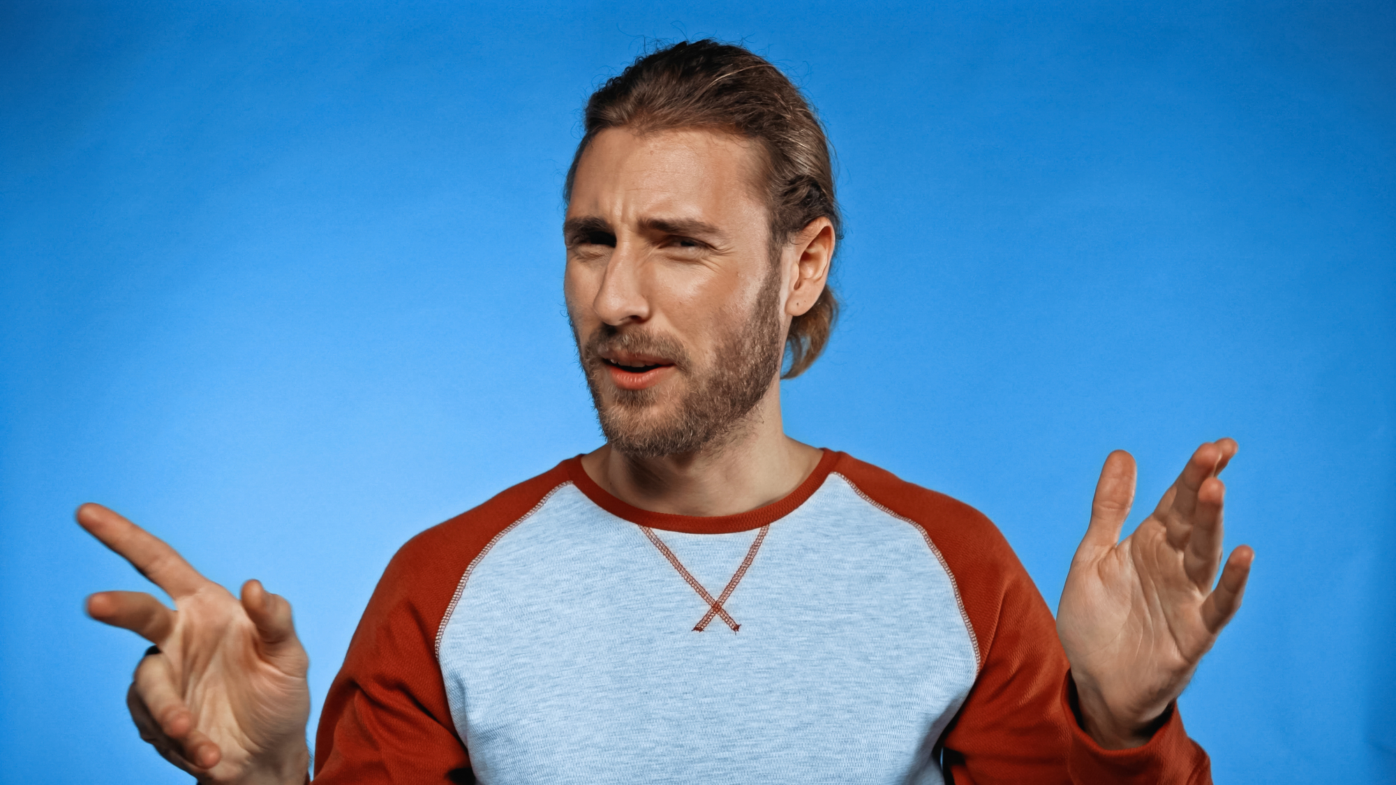 A person with long hair tied back and a beard is making an expressive face, narrowing their eyes and pointing with both hands. They are wearing a light blue longsleeve shirt with red sleeves against a solid blue background.