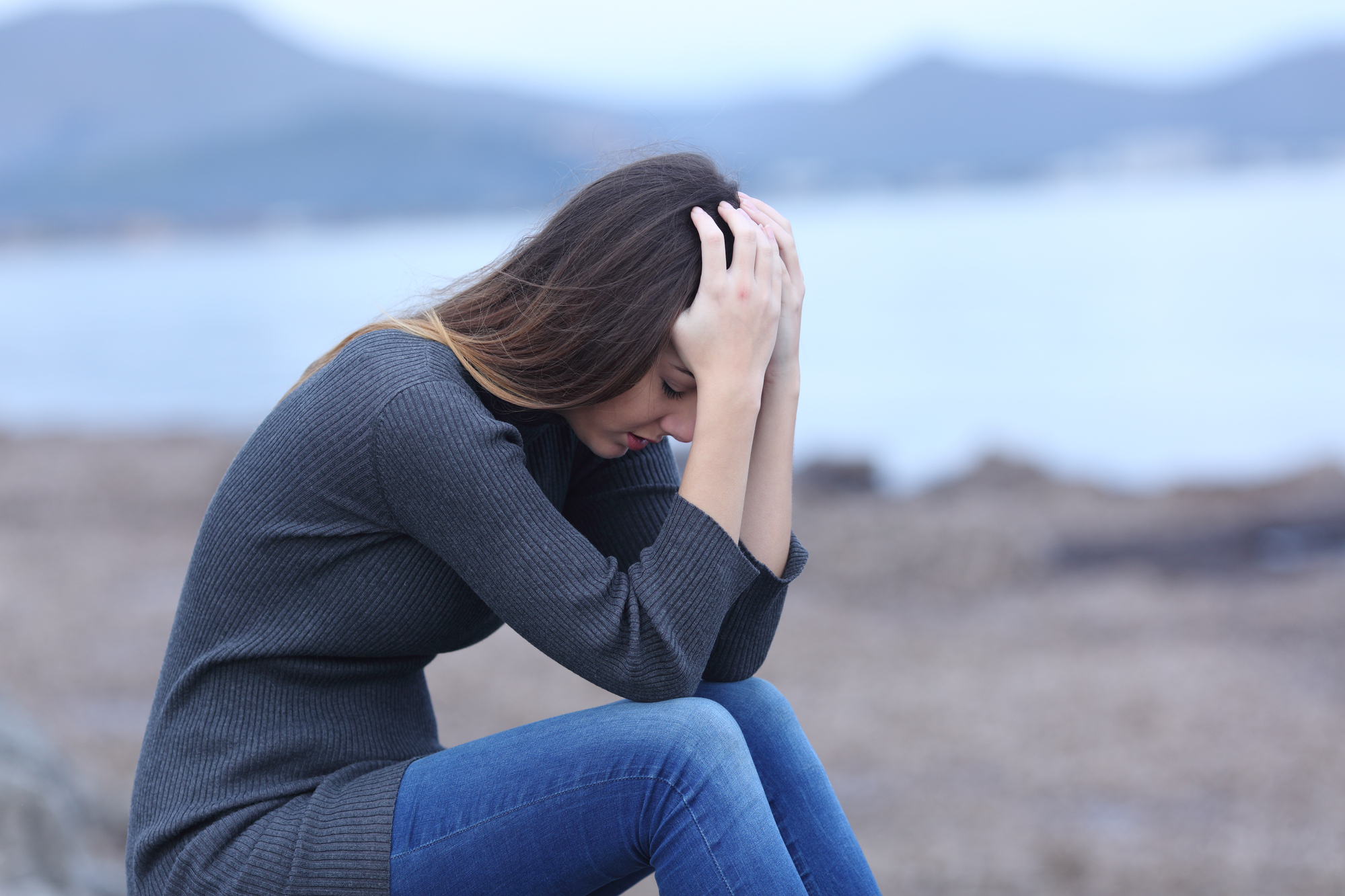A woman with long hair sits outdoors near the water with her head in her hands, appearing distressed. She is wearing a grey sweater and blue jeans. In the background, there's an out-of-focus view of mountains and water.