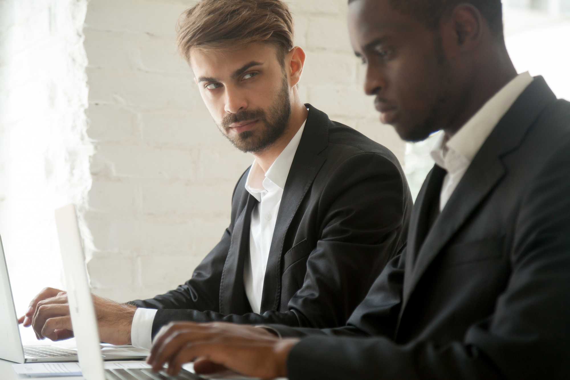 Two men in suits are working together on laptops. The man on the left has a beard and is looking at the man on the right, who is focused on his screen. They are seated at a white desk against a white brick wall background.
