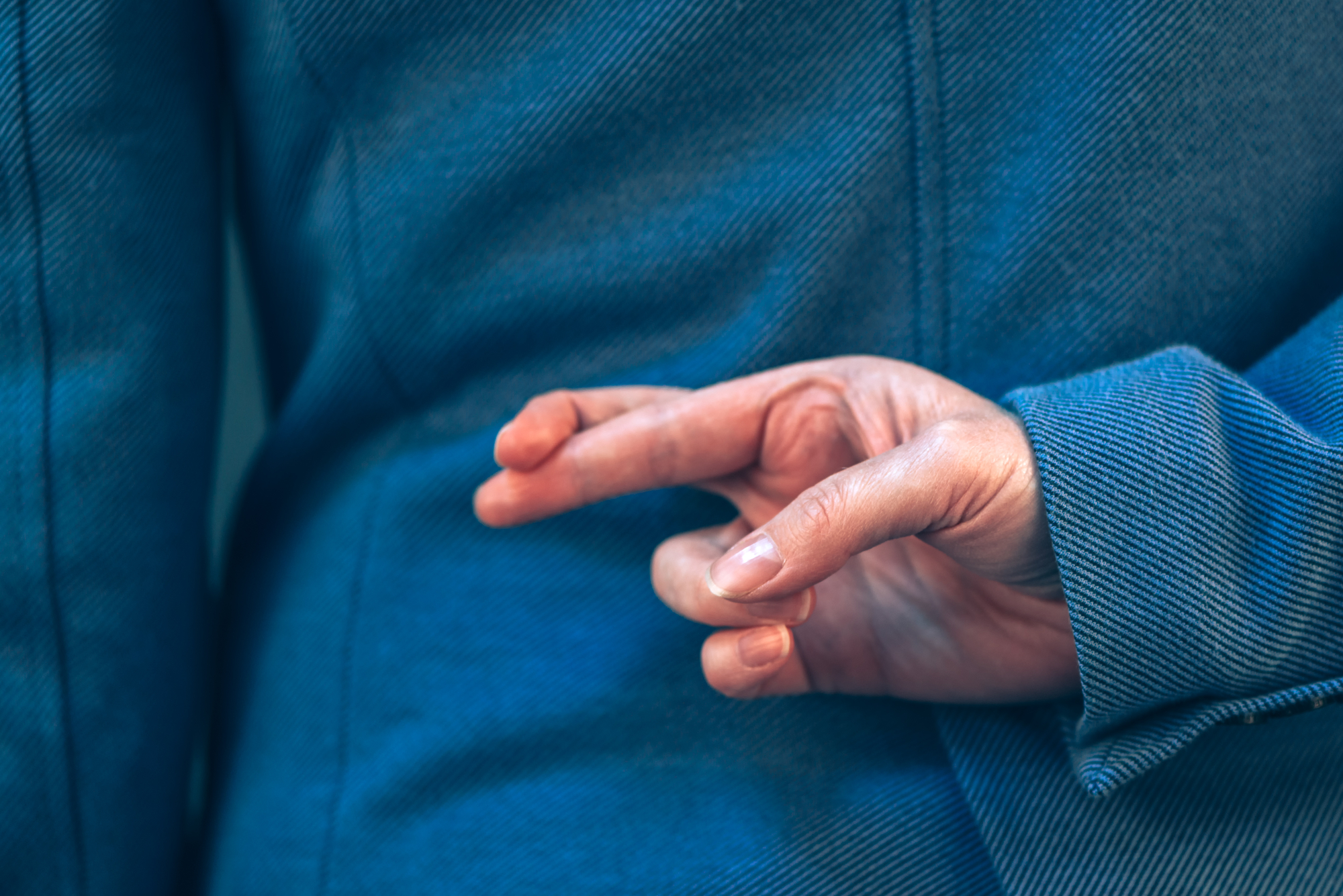 A person wearing a blue jacket is holding their right hand behind their back with their fingers crossed. The gesture is traditionally associated with superstition or deceit. The focus is on the hand and the texture of the jacket.