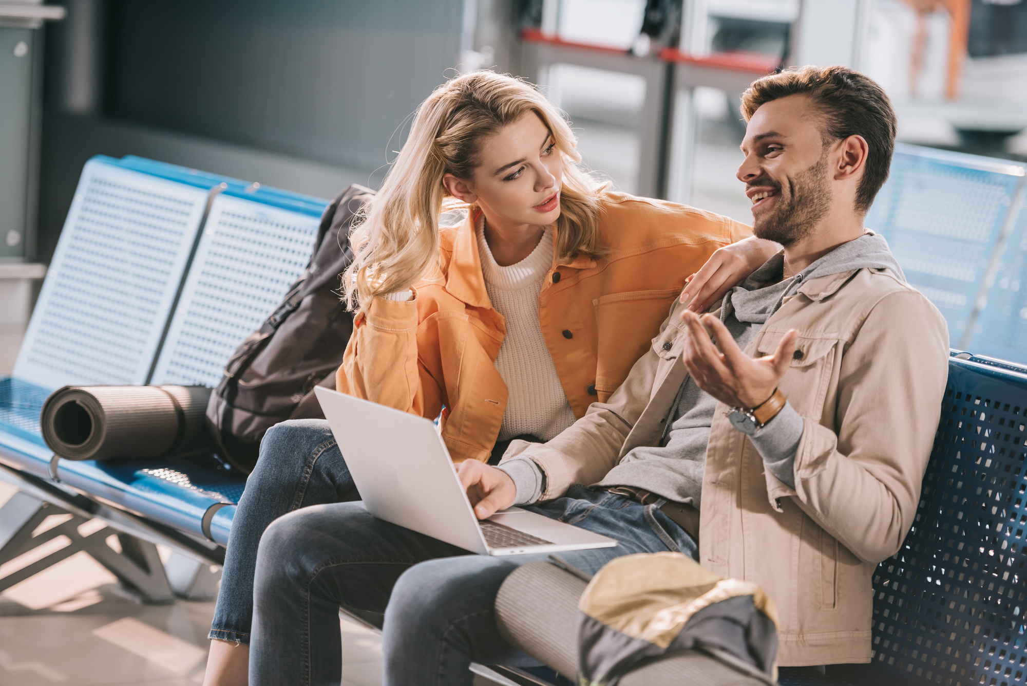 A man and woman sit on blue chairs in an airport, engaged in conversation. The man gestures animatedly while the woman, with her arm around his shoulder, listens attentively. A laptop rests on the man's lap, and a black backpack and yoga mat are nearby.