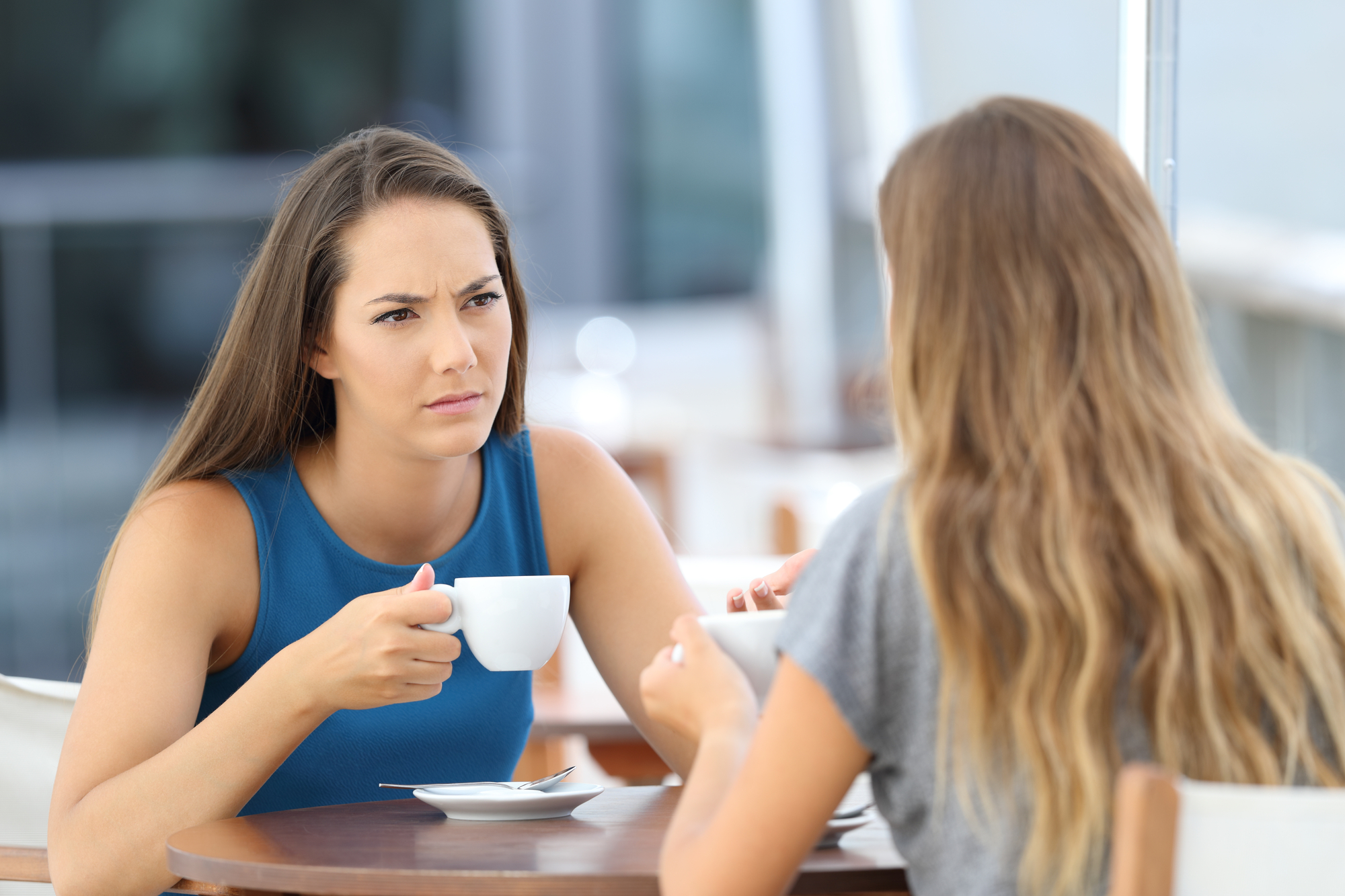 Two women are sitting at a table outdoors, engaged in a serious conversation. The woman on the left is holding a white coffee cup and looking intently at the woman on the right, who has her back to the camera. The setting is casual and appears to be a café.