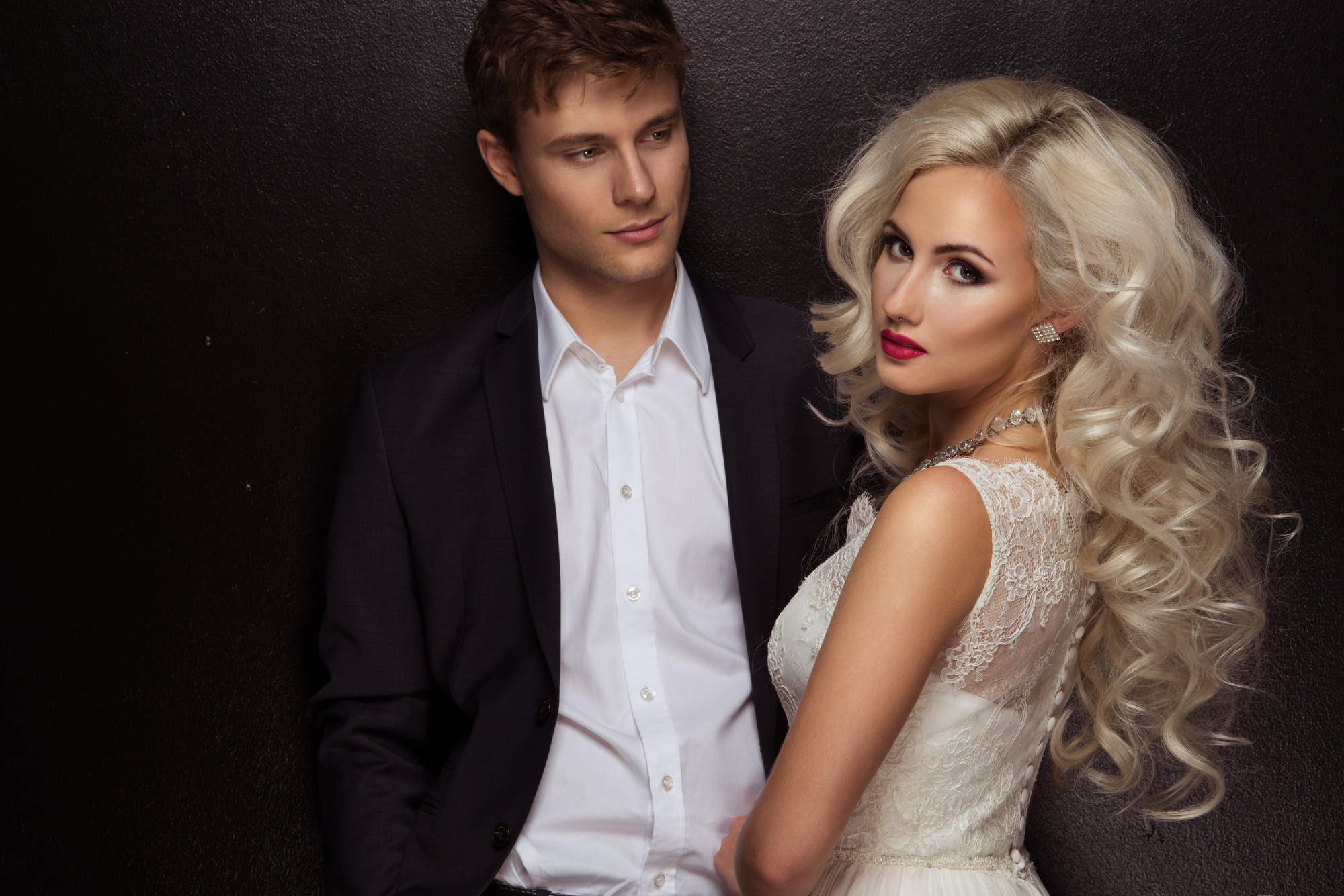 A man and a woman pose together against a black background. The man is wearing a dark suit with a white shirt, and the woman, who has long, wavy blonde hair, is dressed in a white lace dress. Both are looking slightly off camera, with the woman glancing back.
