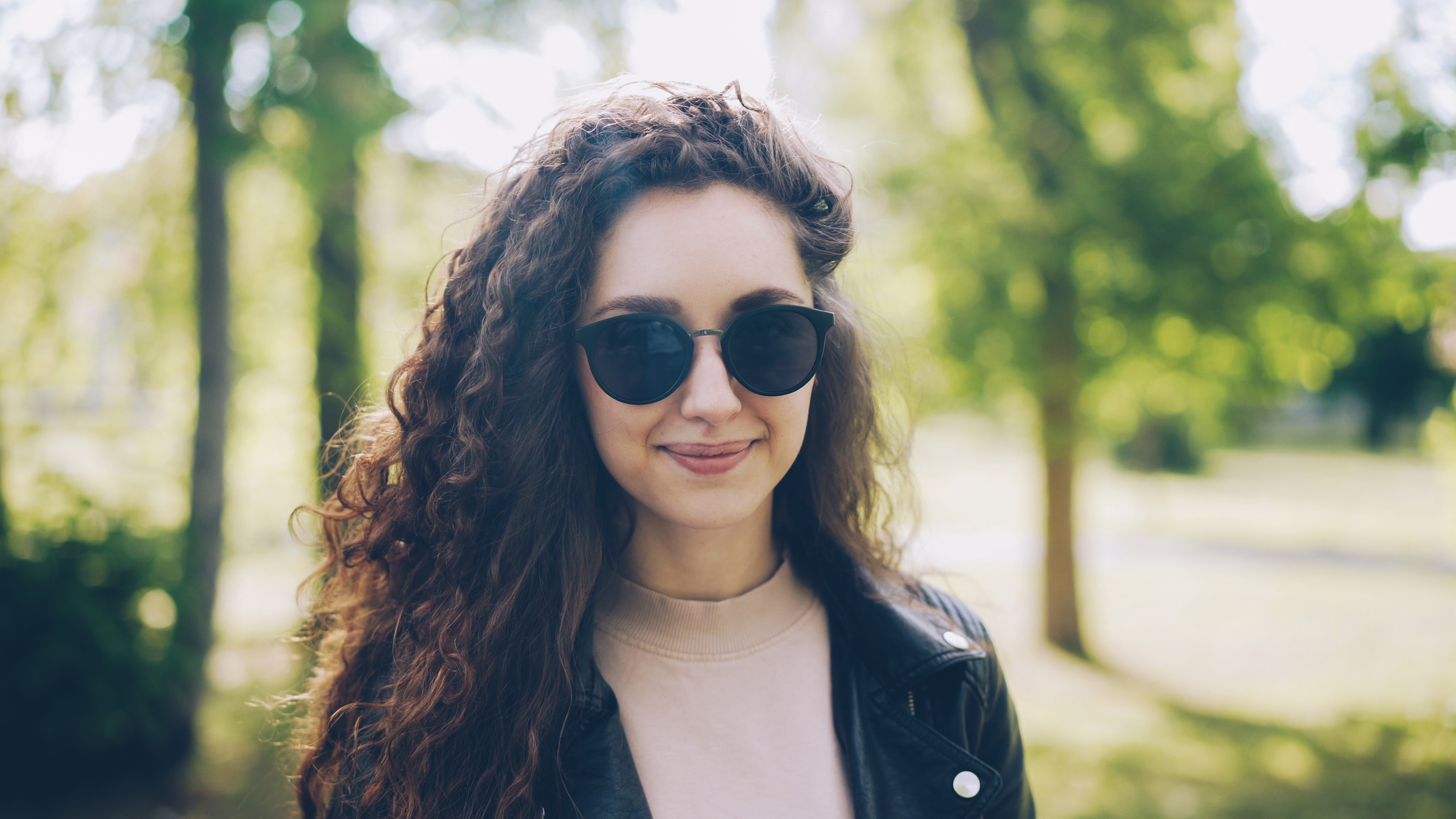 A woman with long curly hair, wearing round sunglasses and a black leather jacket, is standing outdoors in a sunlit park with green foliage in the background, smiling gently at the camera.